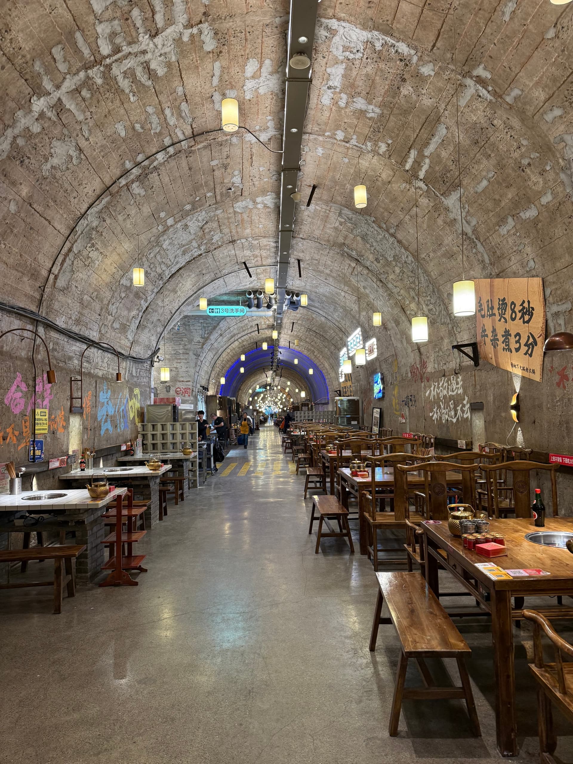 Ancien abri anti-aérien de Chongqing reconverti en restaurant : long tunnel voûté en béton, tables et tabourets de bois alignés le long des parois, lanternes suspendues