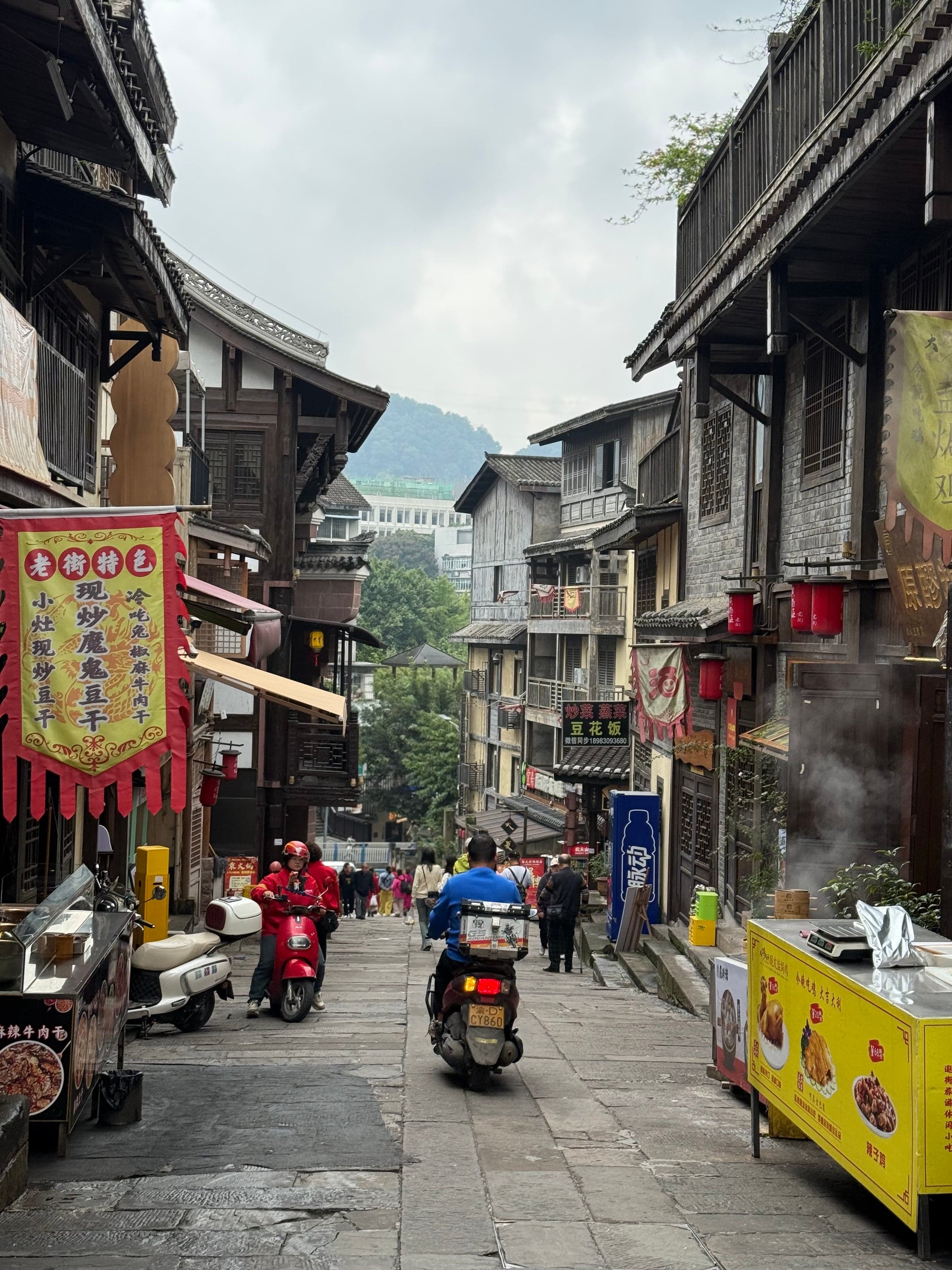 Rue pavée de Ciqikou avec maisons en bois, scooters et échoppes de street food sous un ciel couvert