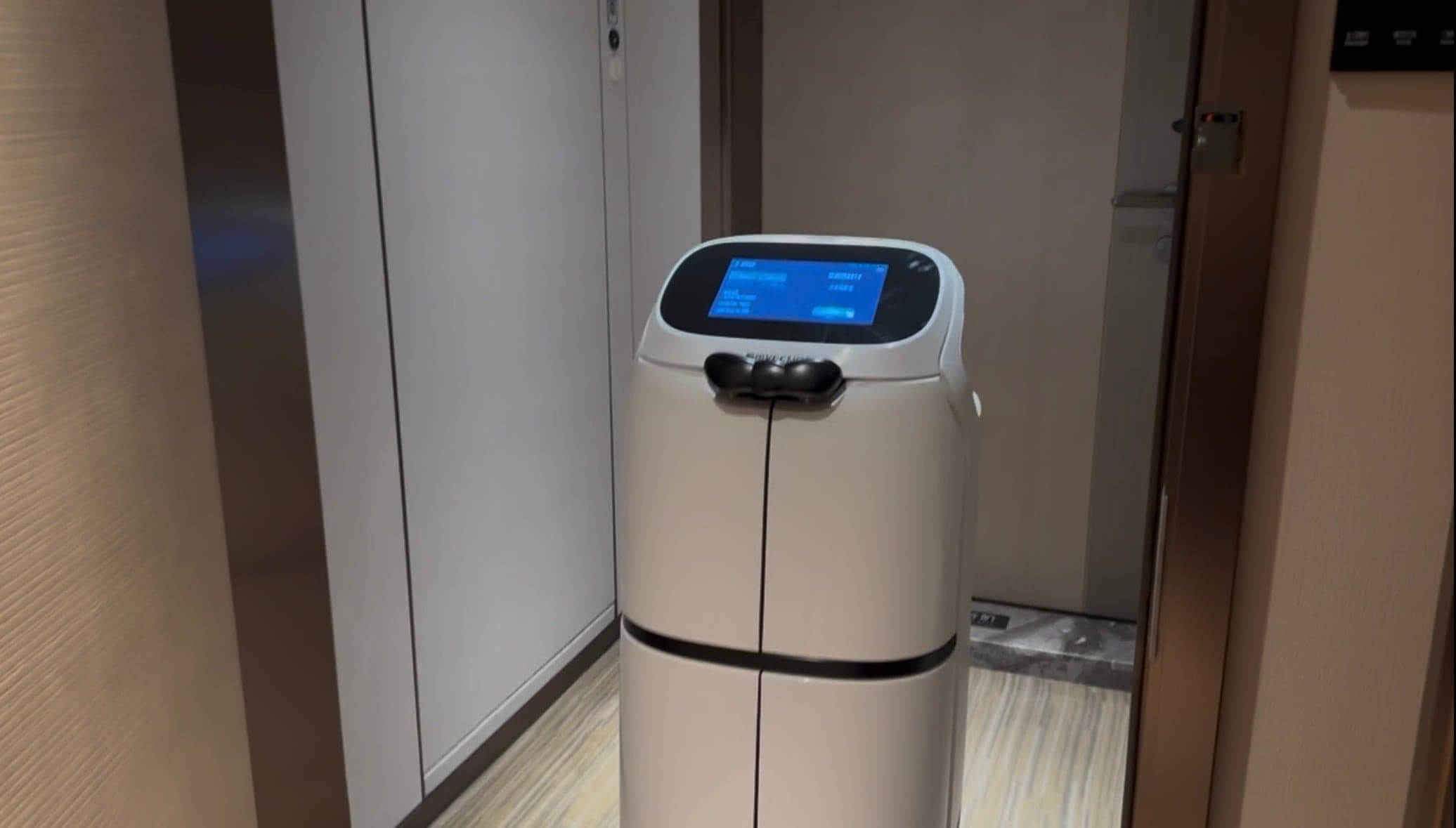 Robot livreur blanc à écran tactile dans le couloir d'un hôtel chinois, arrêté devant les portes d'ascenseur, coffre fermé contenant une commande