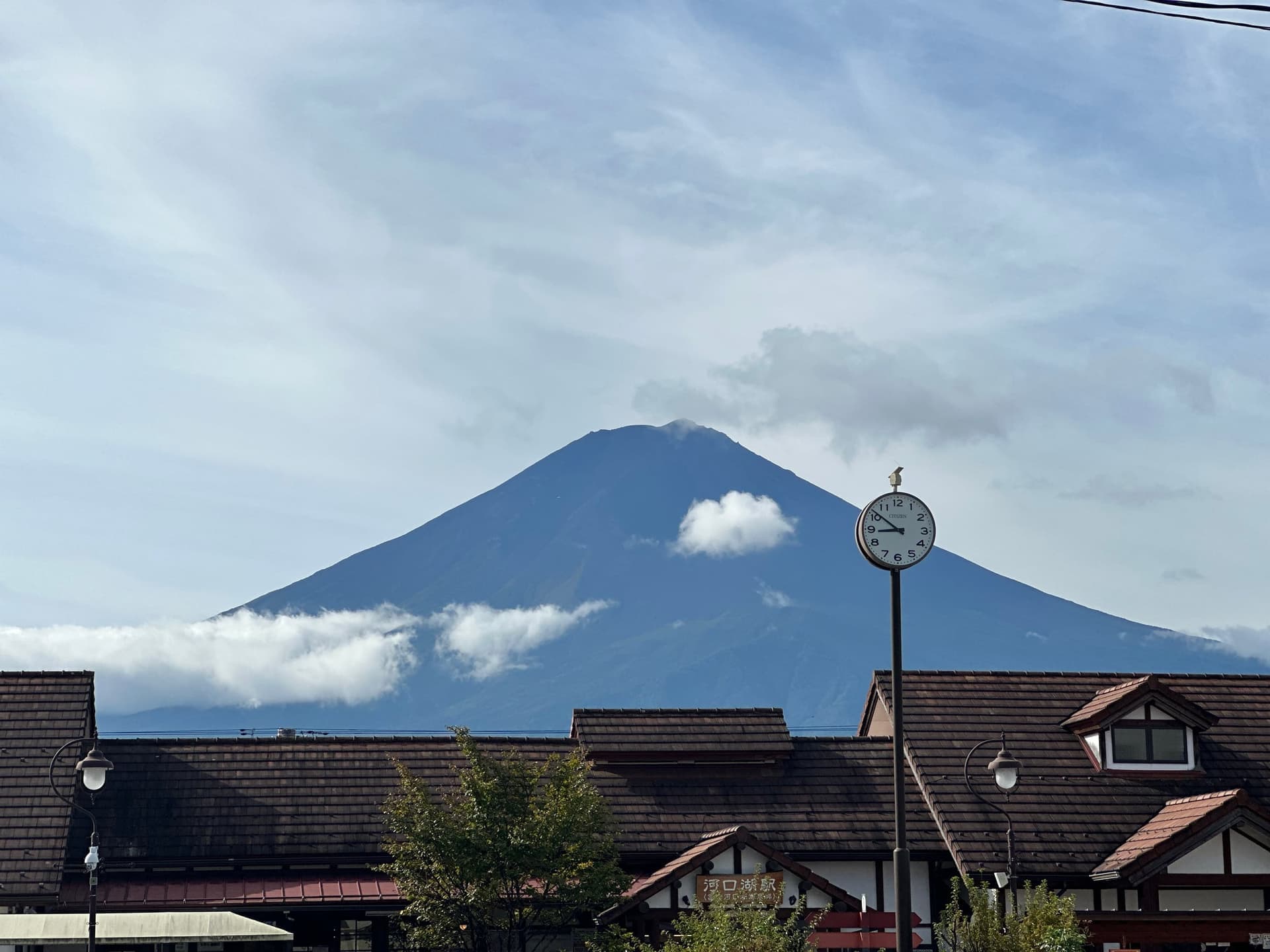 Le mont Fuji dégagé au-dessus d'une ceinture de nuages, vu depuis Kawaguchiko, toits d'une gare au premier plan avec une horloge