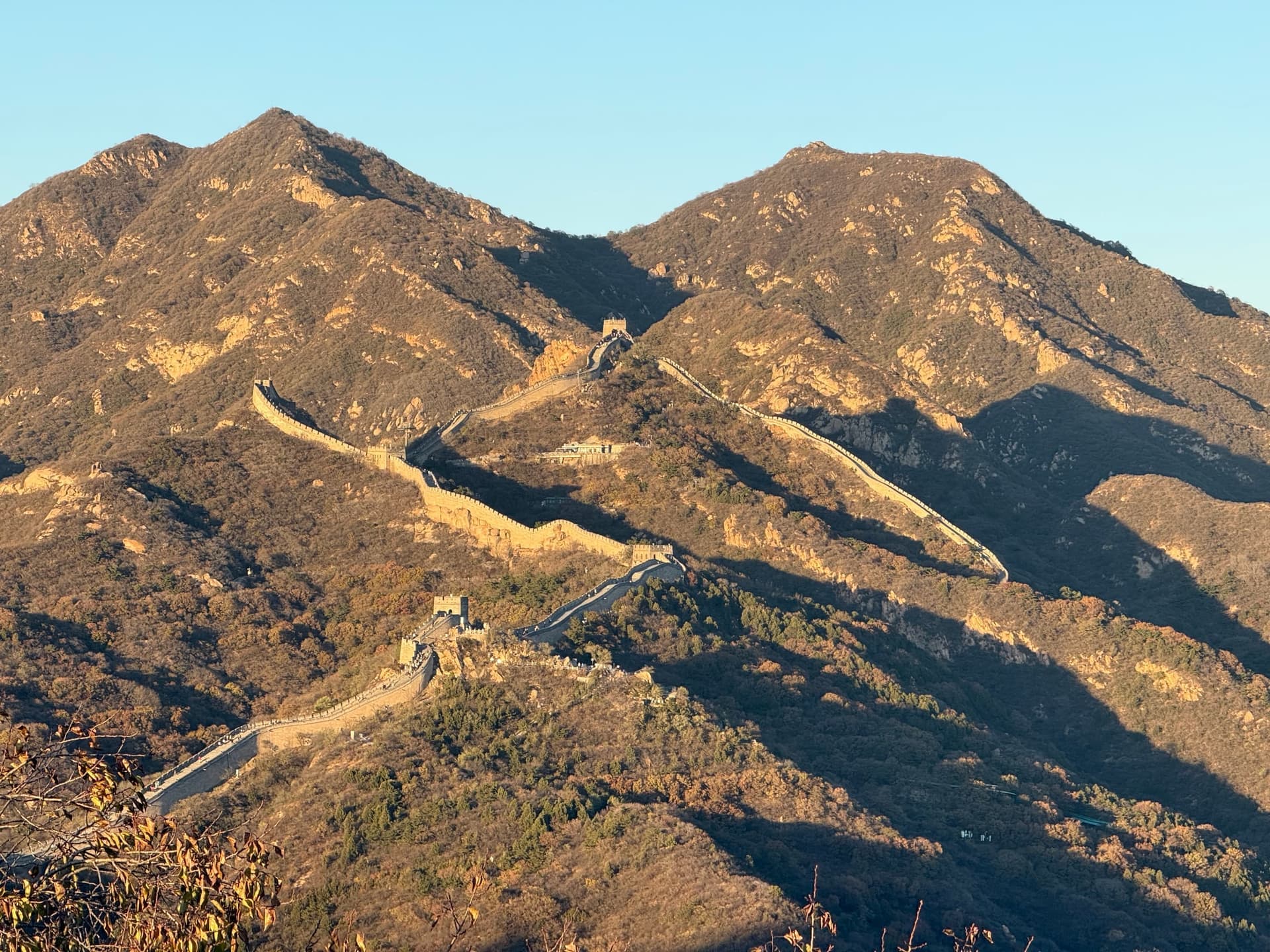 Vue panoramique de la Grande Muraille à Badaling serpentant sur des crêtes montagneuses successives, ciel clair, végétation automnale dorée et brune, tours de garde visibles le long de la muraille