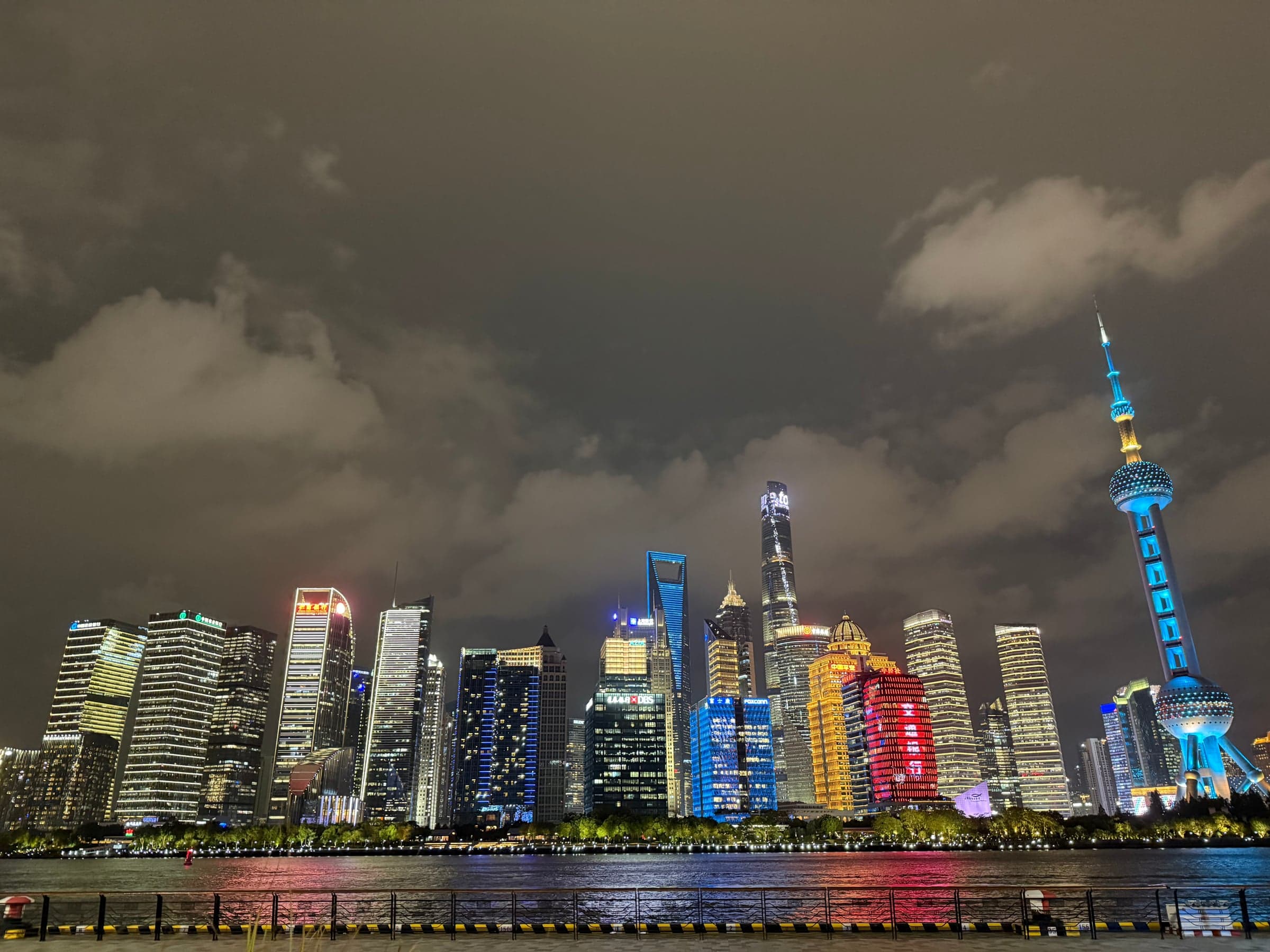 Panorama nocturne des tours de Pudong depuis la promenade du Bund, façades illuminées de couleurs changeantes sous un ciel couvert