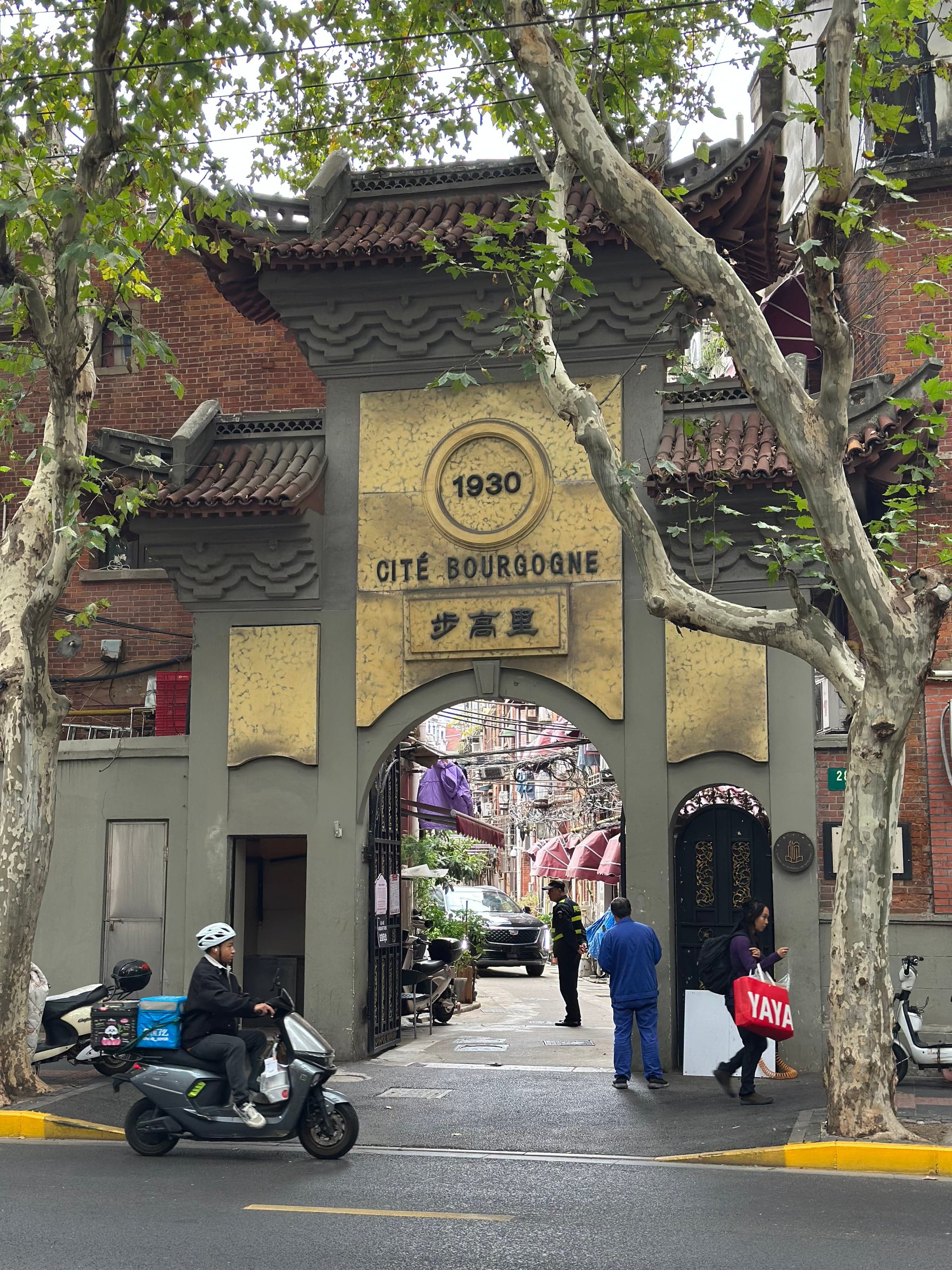 Porte d'entrée en pierre de la Cité Bourgogne à Shanghai, datée 1930, arche donnant sur une ruelle d'habitation bordée de platanes, passants et scooter de livraison