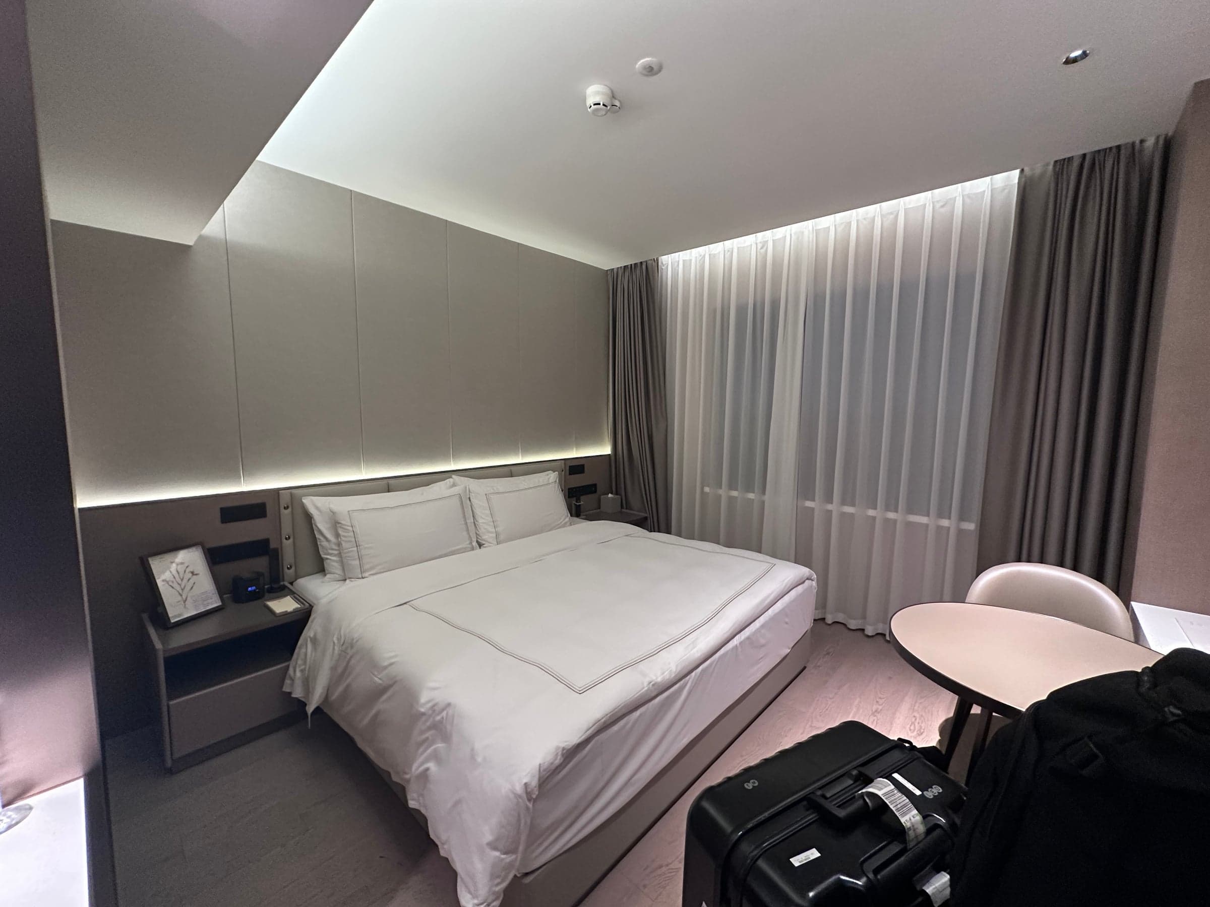 Chambre d'hôtel moderne à Shanghai, grand lit blanc, tête de lit rétroéclairée, rideaux jusqu'au sol et valise posée près du bureau