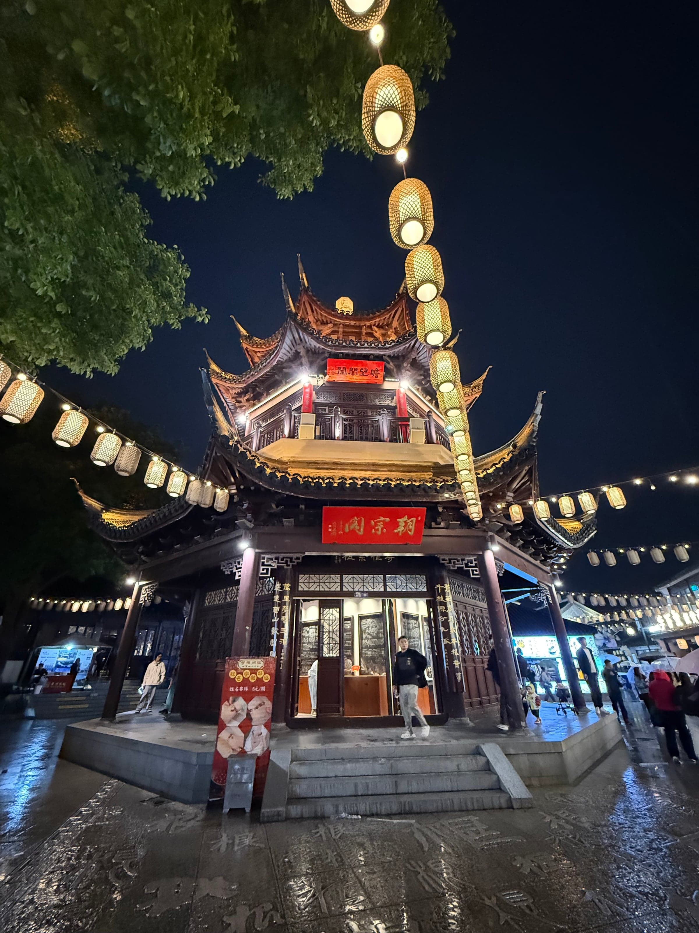 Pavillon de bois à deux niveaux illuminé la nuit sur le canal Shantang à Suzhou, guirlandes de lanternes tendues au-dessus de la rue et pavés mouillés