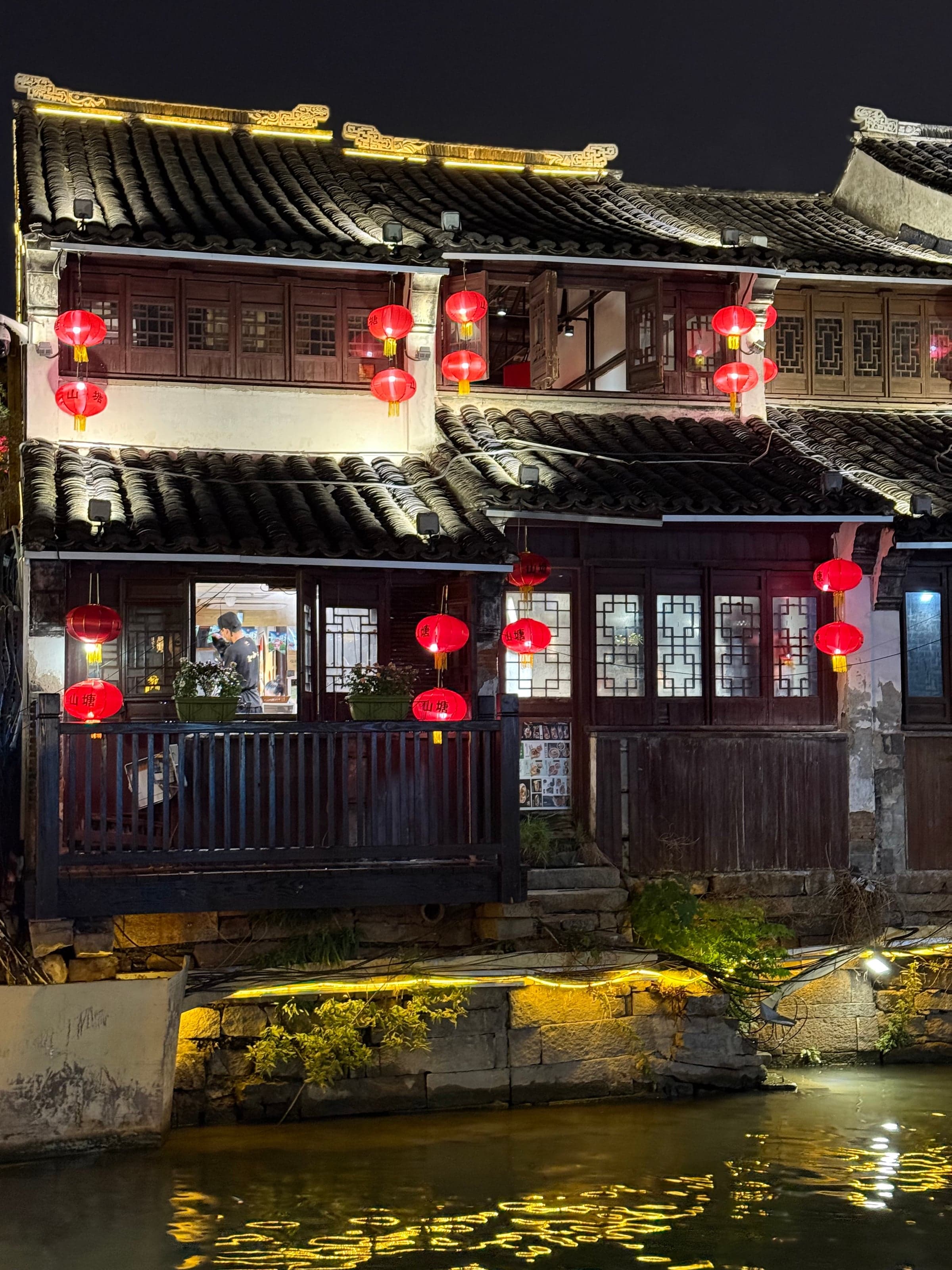 Maisons anciennes en bois au bord du canal Shantang à Suzhou la nuit, lanternes rouges allumées le long des façades, reflets dorés sur l'eau