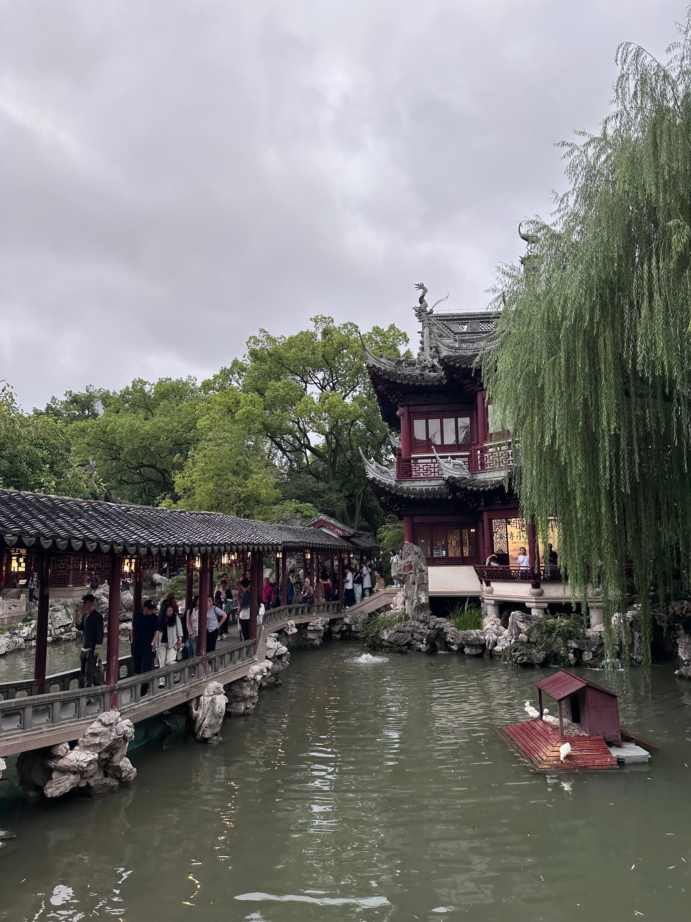 Galerie couverte longeant l'étang du jardin Yu, visiteurs alignés sous le toit, saules et barque rouge sur l'eau, ciel gris