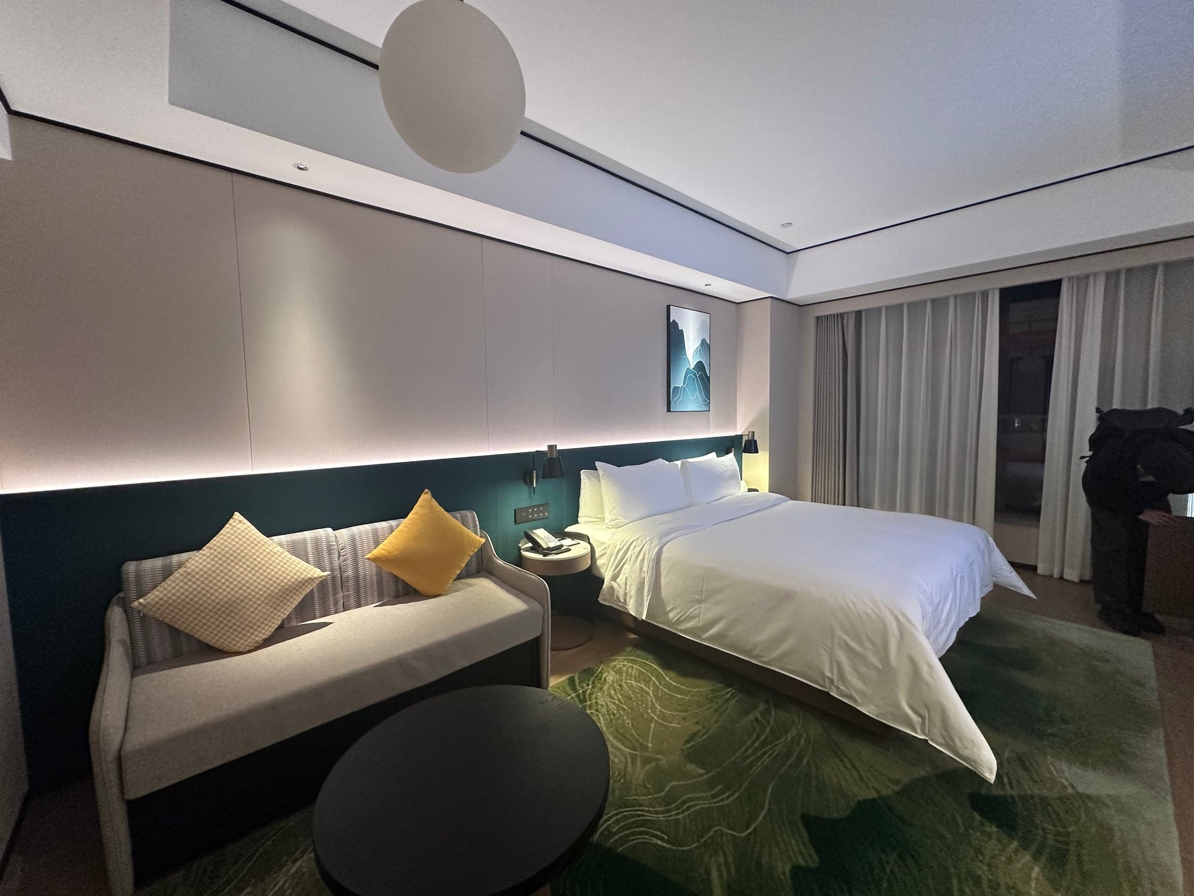 Chambre d'hôtel spacieuse en Chine : grand lit blanc, canapé et coussins jaunes, tapis vert, éclairage tamisé et estampe de montagne au mur