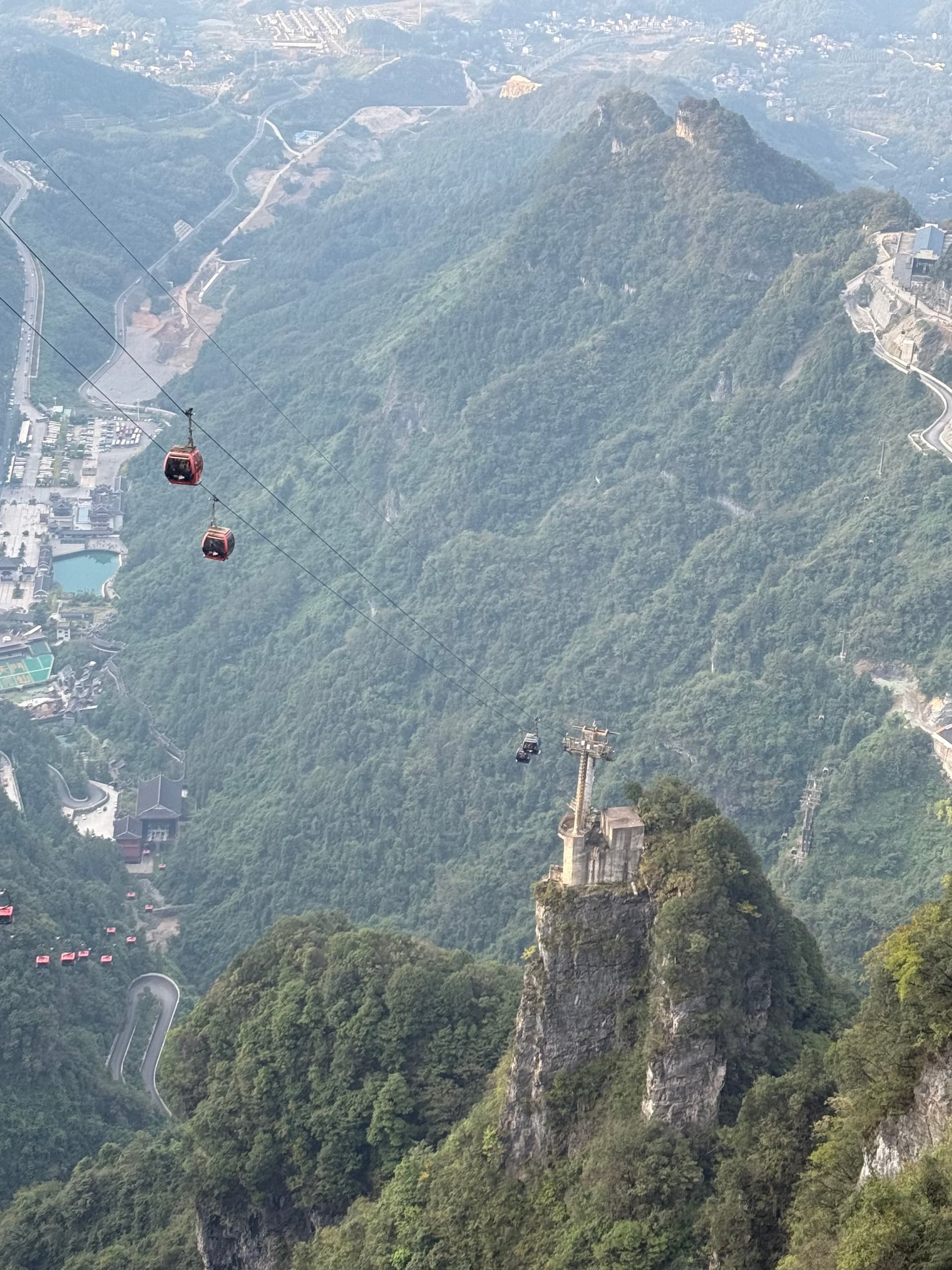 Vue plongeante depuis une cabine de téléphérique sur une vallée boisée, câbles et cabines rouges de la télécabine, éperon rocheux coiffé d'un pavillon et ville en contrebas