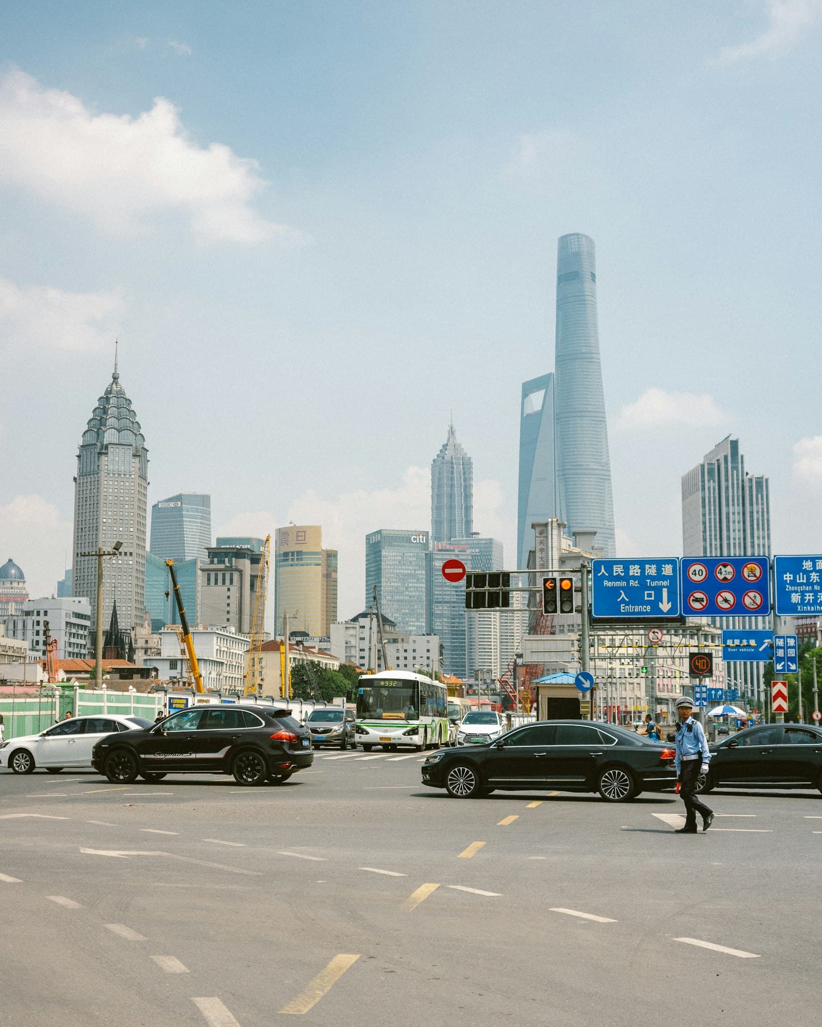 Carrefour de Shanghai en journée, berlines et bus à l'arrêt devant un agent de circulation en uniforme bleu, panneaux routiers bilingues et gratte-ciel de Pudong dont la Shanghai Tower en arrière-plan