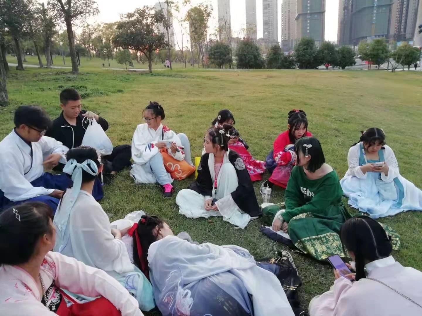 Un groupe de jeunes Chinois en hanfu assis dans l'herbe d'un parc urbain, tours d'habitation à l'arrière-plan