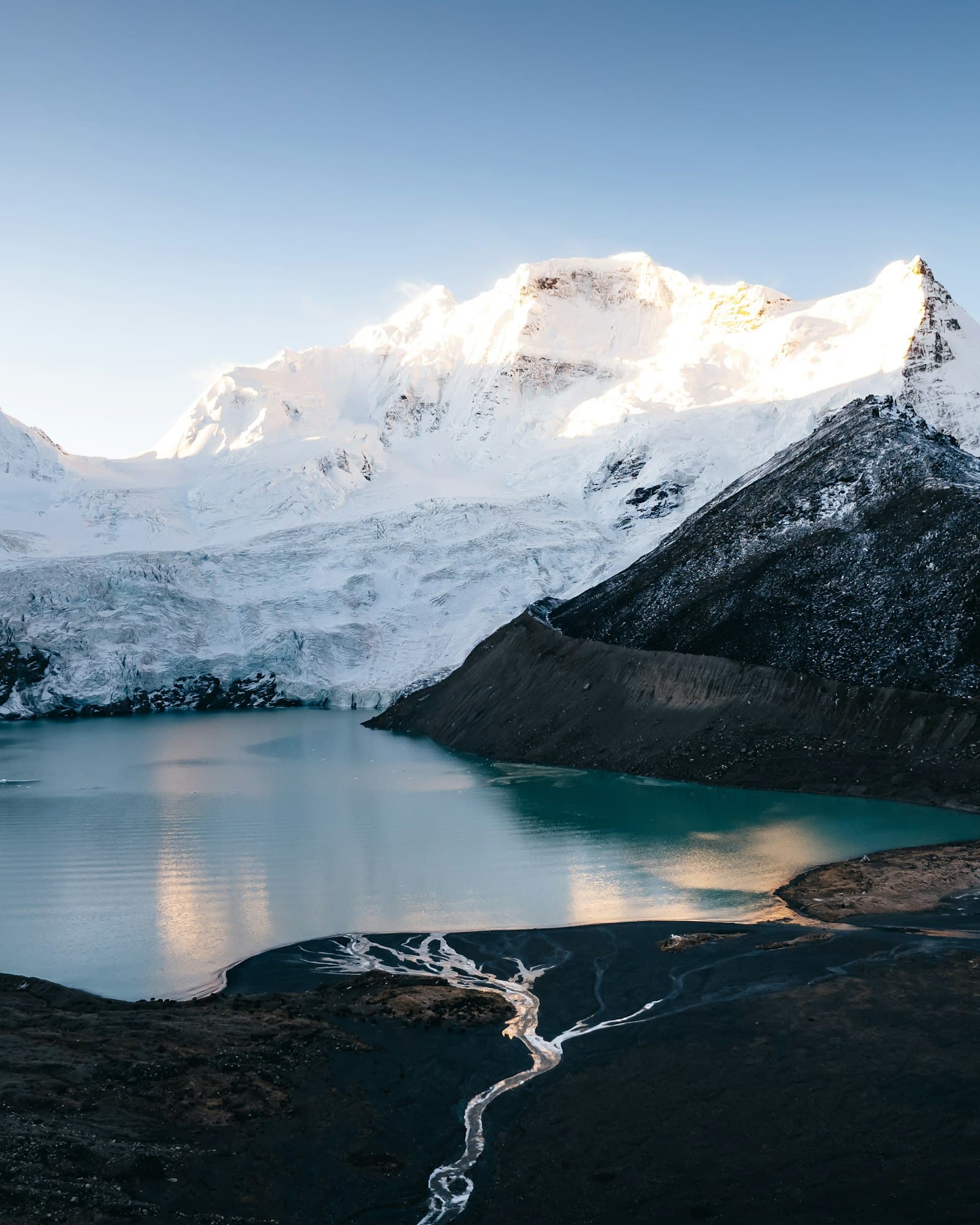 Lac glaciaire aux eaux turquoise sur le plateau tibétain au lever du soleil, langue de glacier plongeant dans l'eau et sommet enneigé éclairé en arrière-plan