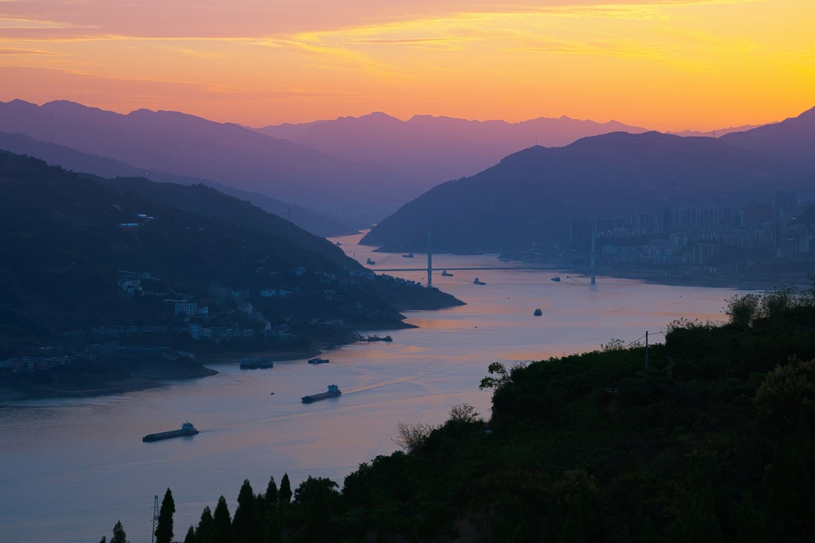 Le Yangtsé au coucher du soleil près de Fengjie : le fleuve large parcouru de barges, un pont à haubans, et la ville moderne construite à flanc de colline avant l'entrée des gorges
