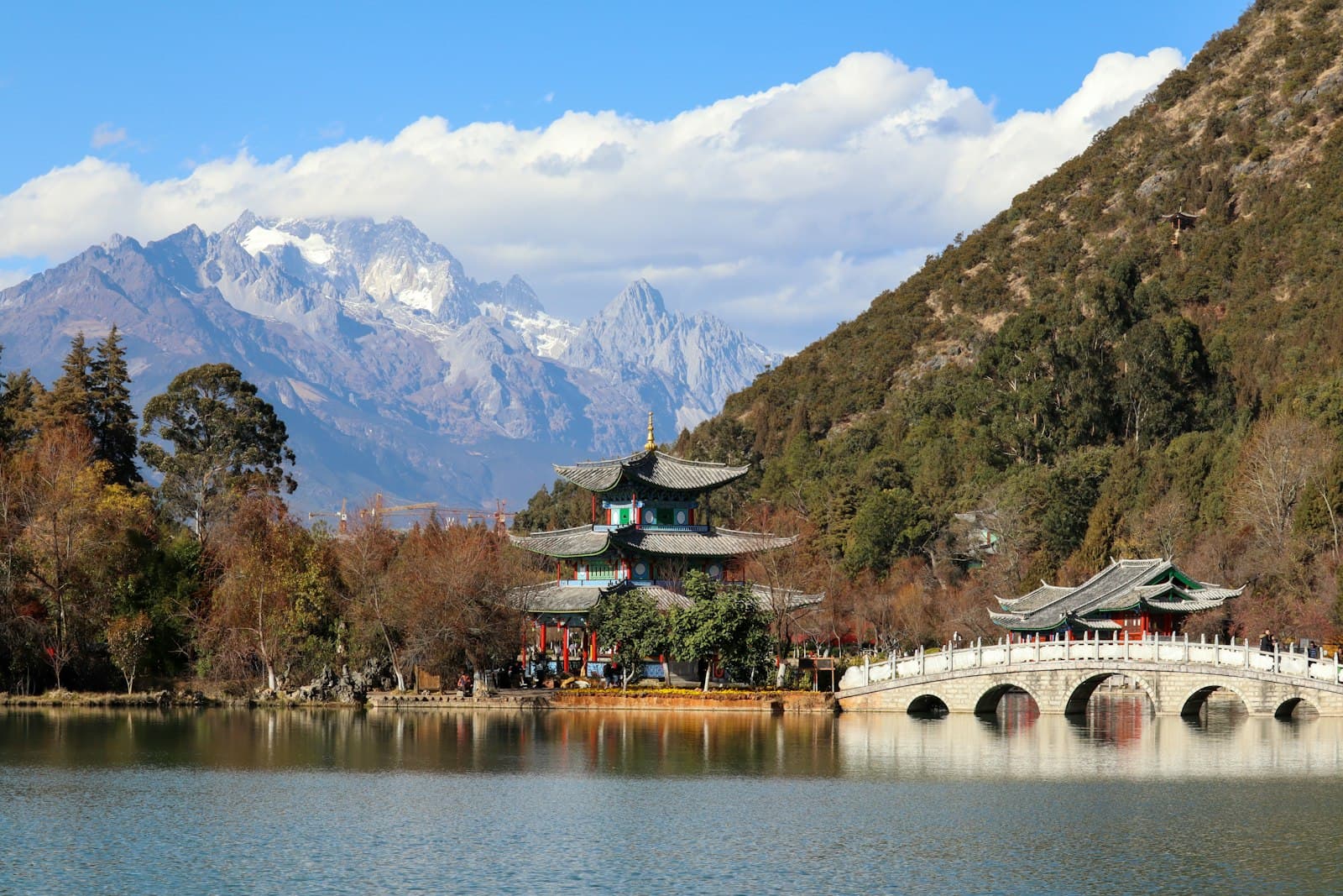 L'étang du Dragon Noir à Lijiang, un pavillon traditionnel et un pont de pierre en arche sur l'eau calme, la montagne enneigée du Dragon de Jade en arrière-plan sous un ciel bleu