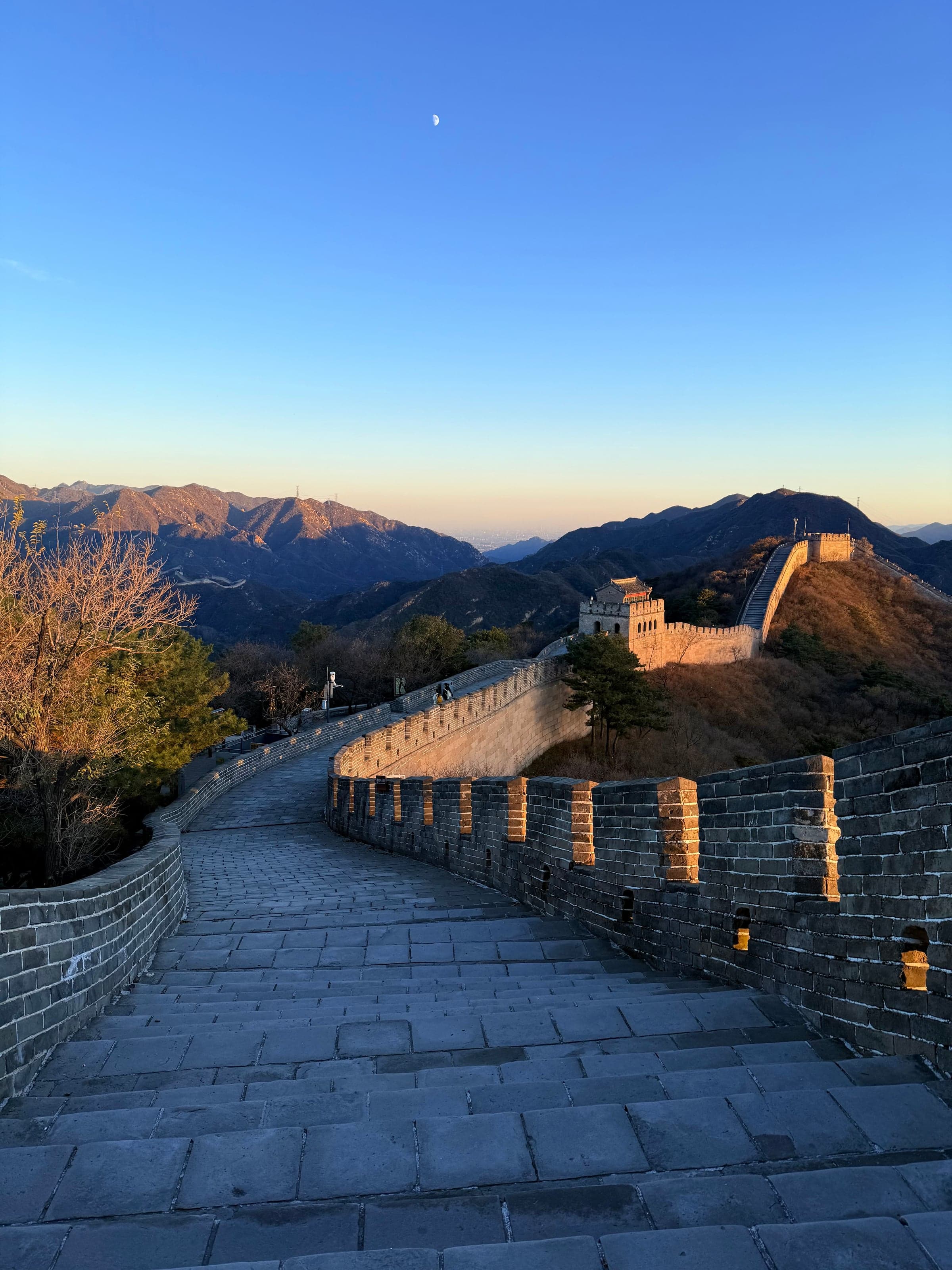 La Grande Muraille déserte à Badaling au coucher du soleil, parapets crénelés en ombre et muraille dorée serpentant vers les montagnes, lune visible