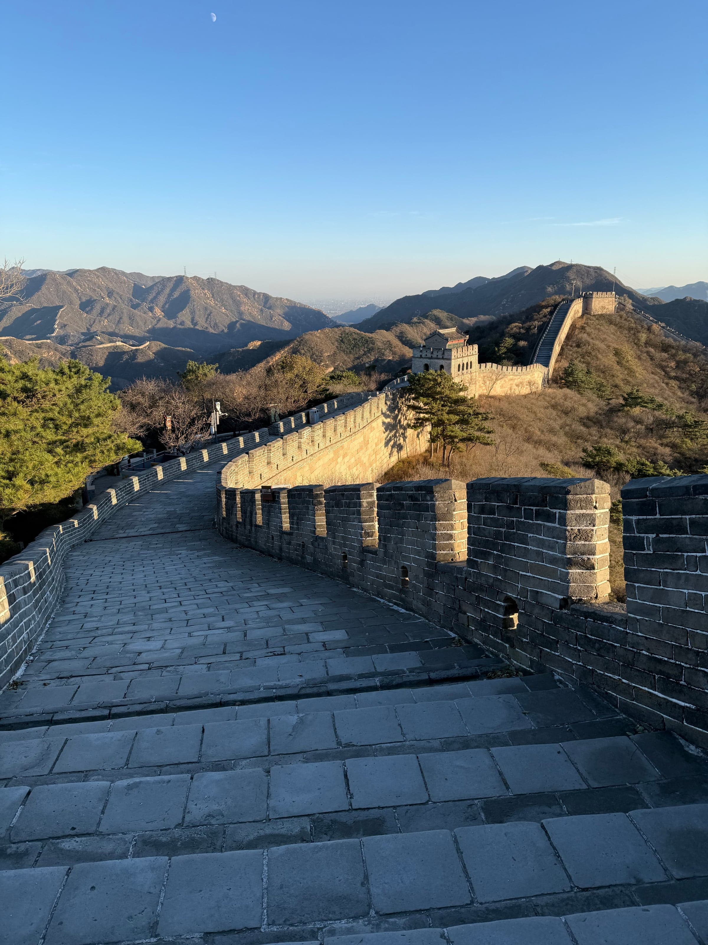 Chemin de ronde de la Grande Muraille à Badaling entièrement vide dans la lumière dorée du soir, tour de guet et crêtes au fond, lune pâle dans le ciel
