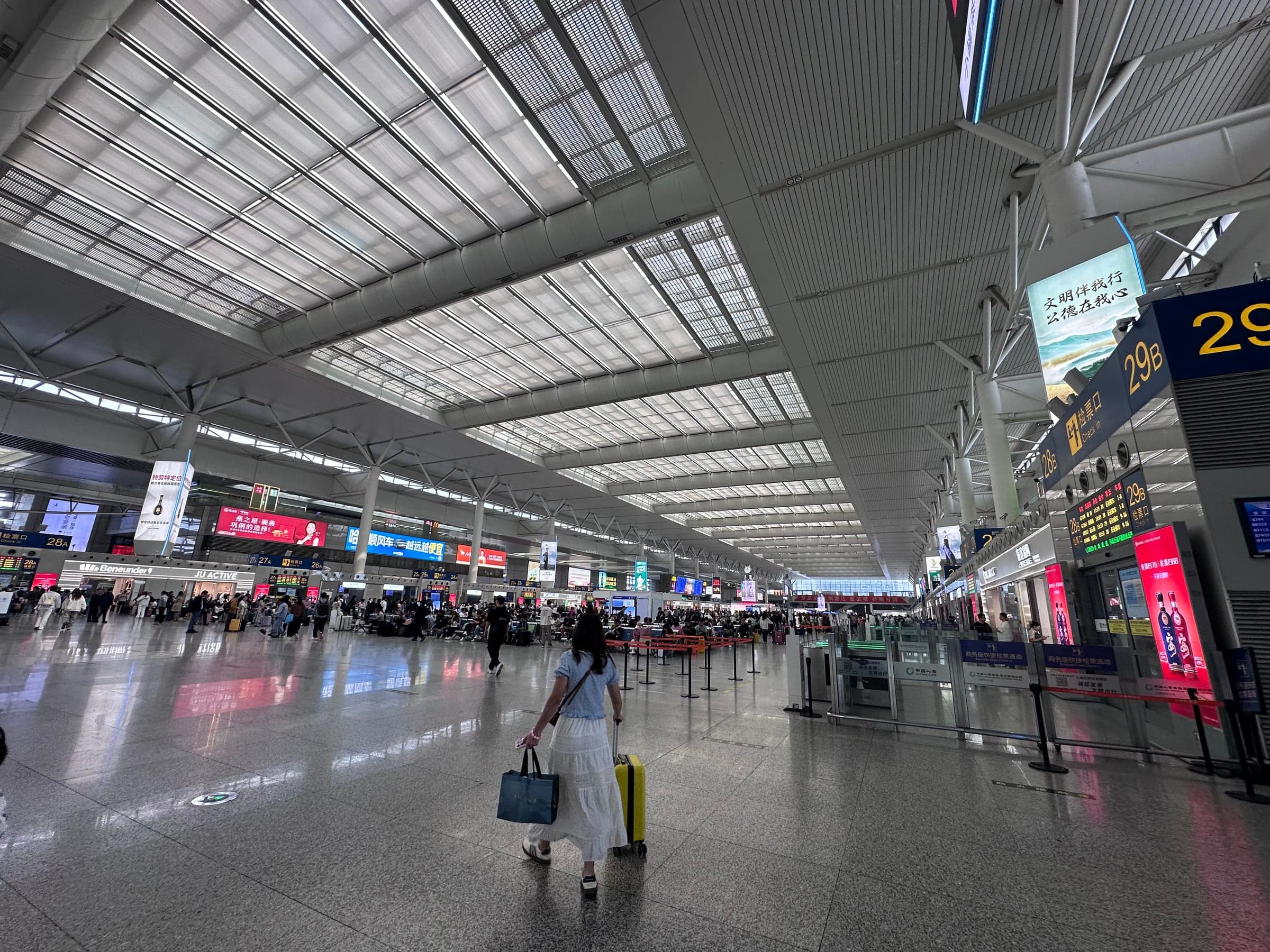 Immense hall de la gare de Shanghai Hongqiao sous sa verrière, voyageuse tirant une valise jaune vers les portiques d'embarquement, écrans et enseignes lumineuses