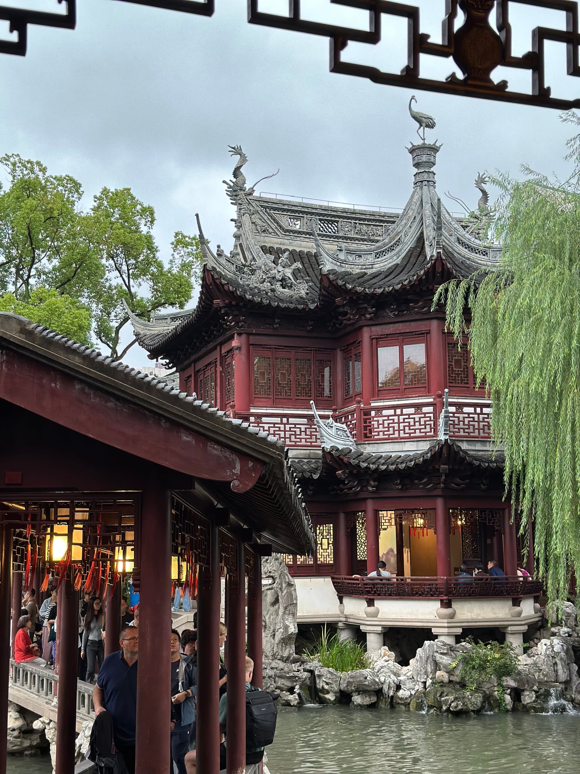 Pavillon à deux niveaux du jardin Yu surplombant un étang, boiseries sombres, saule pleureur et rocaille, visiteurs sous la galerie