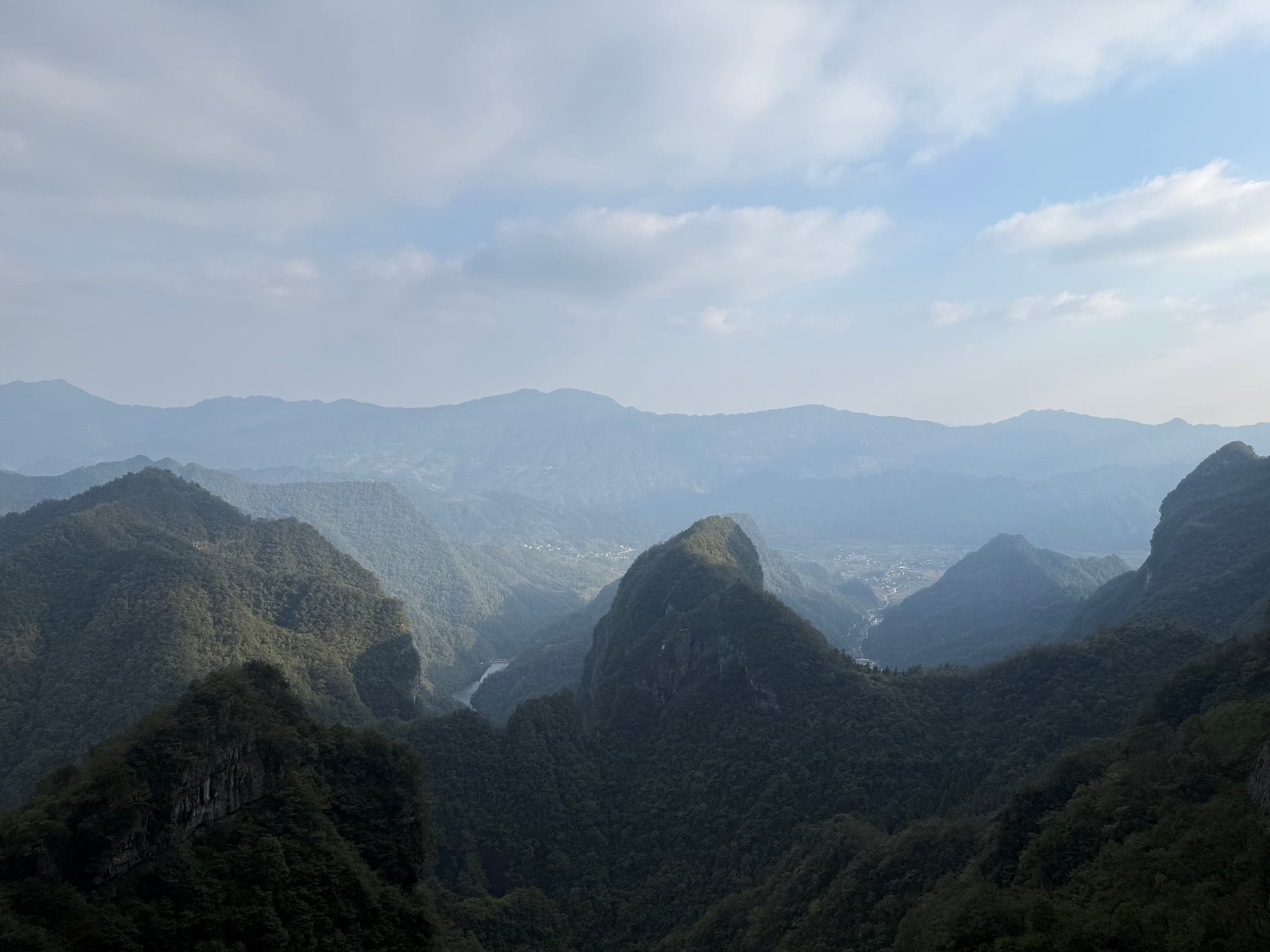 Vue plongeante sur une vallée boisée depuis un point haut de Zhangjiajie, lignes de montagnes bleutées se succédant jusqu'à l'horizon sous des nuages