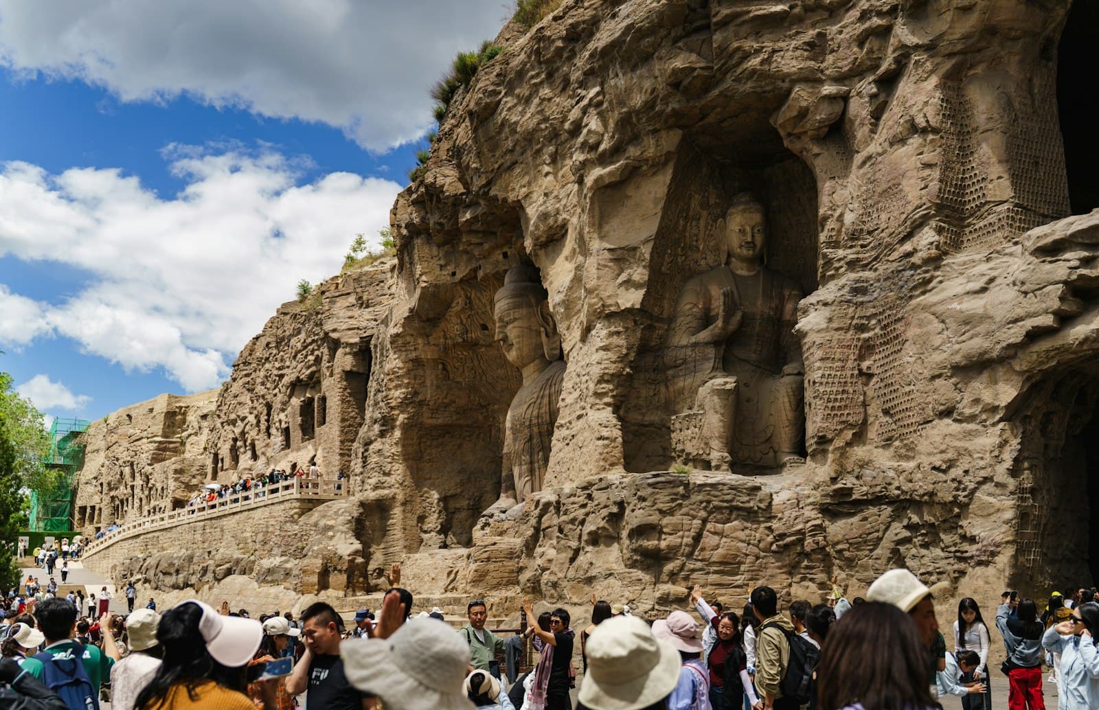 Falaise de grès des grottes de Yungang à Datong : un bouddha colossal assis sculpté dans une niche, la paroi criblée de petites niches, une passerelle de visiteurs et une foule au pied du rocher