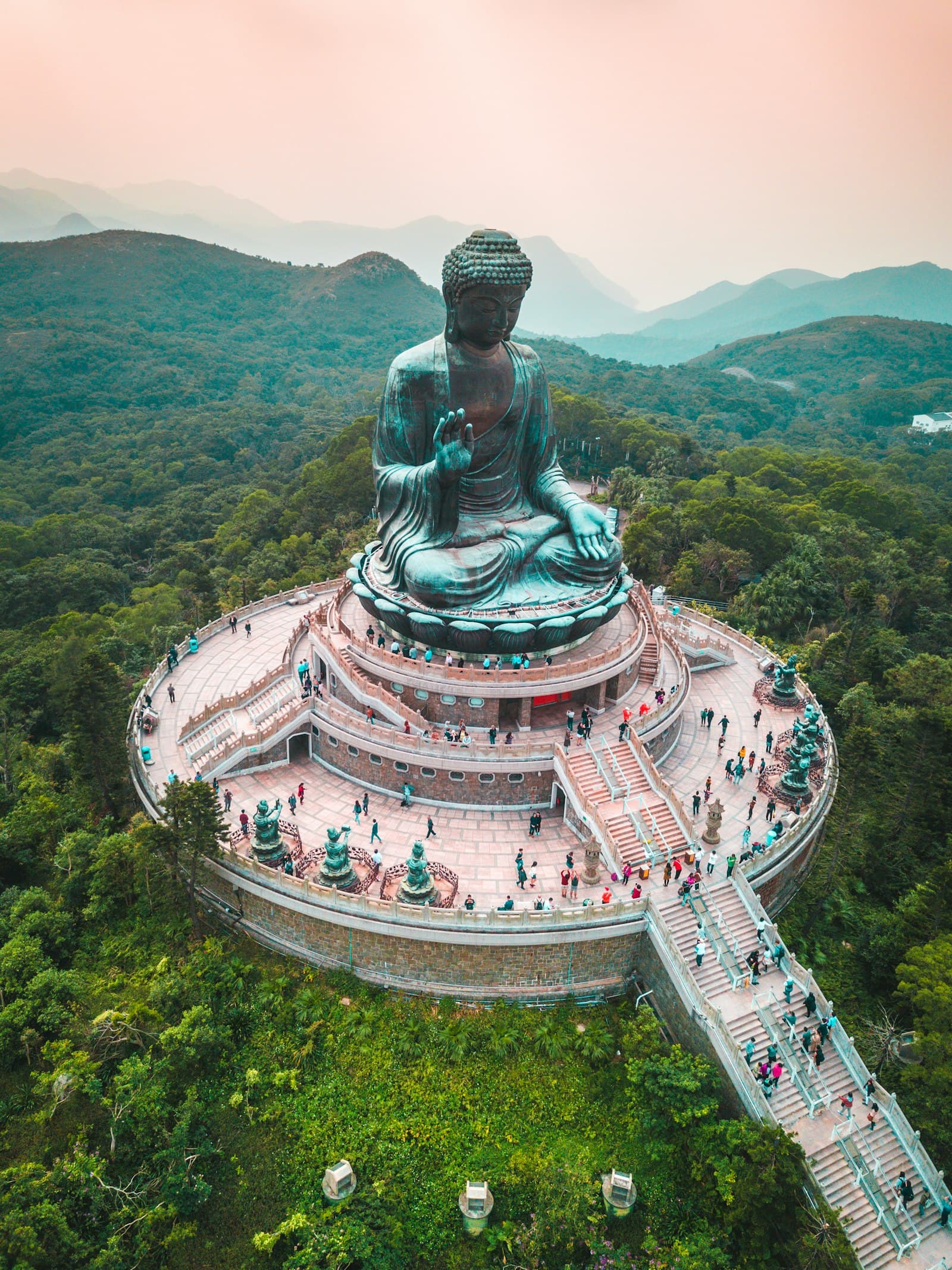 Vue aérienne du Tian Tan Buddha, statue de bronze assise au sommet d'une plateforme circulaire à Lantau, avec l'escalier d'accès couvert de visiteurs et les collines boisées de l'île à l'arrière-plan