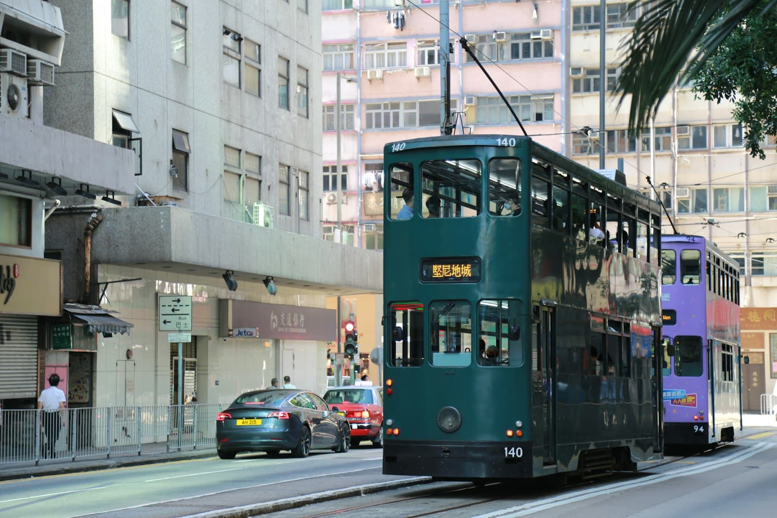 Deux tramways à impériale de Hong Kong dans une rue de l'île, le premier vert foncé portant le numéro 140 et la destination Kennedy Town en caractères chinois, le second violet, entre immeubles anciens et taxi rouge