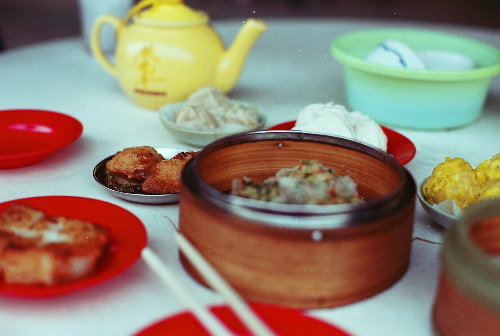 Table de yum cha à Hong Kong avec un panier vapeur en bambou garni de siu mai, une théière jaune, des assiettes rouges de pâté de navet grillé et de beignets, et des baguettes posées au premier plan