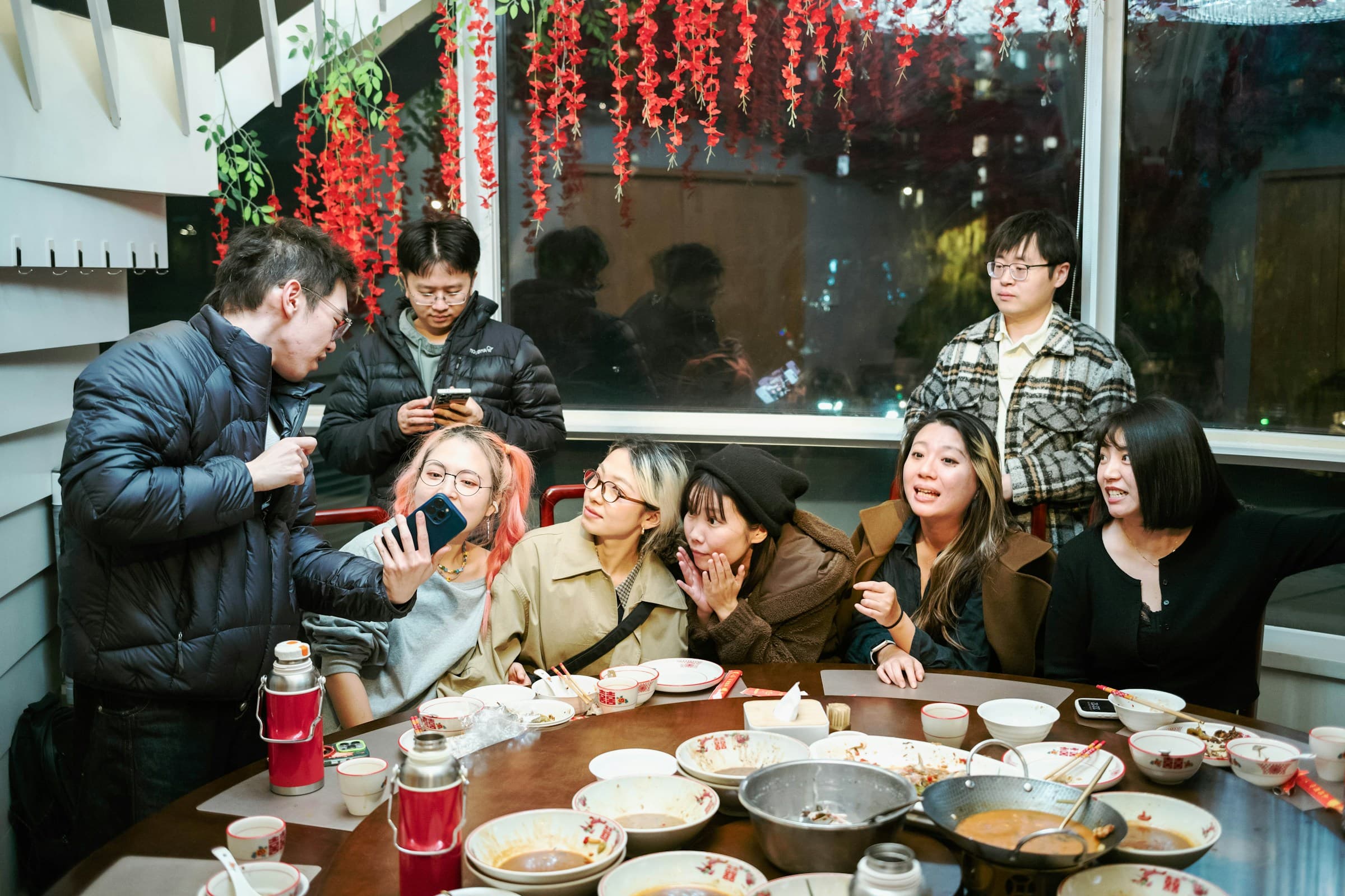 Jeunes convives réunis autour d'une table ronde en fin de repas de Nouvel An chinois, bols au double bonheur, thermos rouges et enveloppes rouges posées sur la table