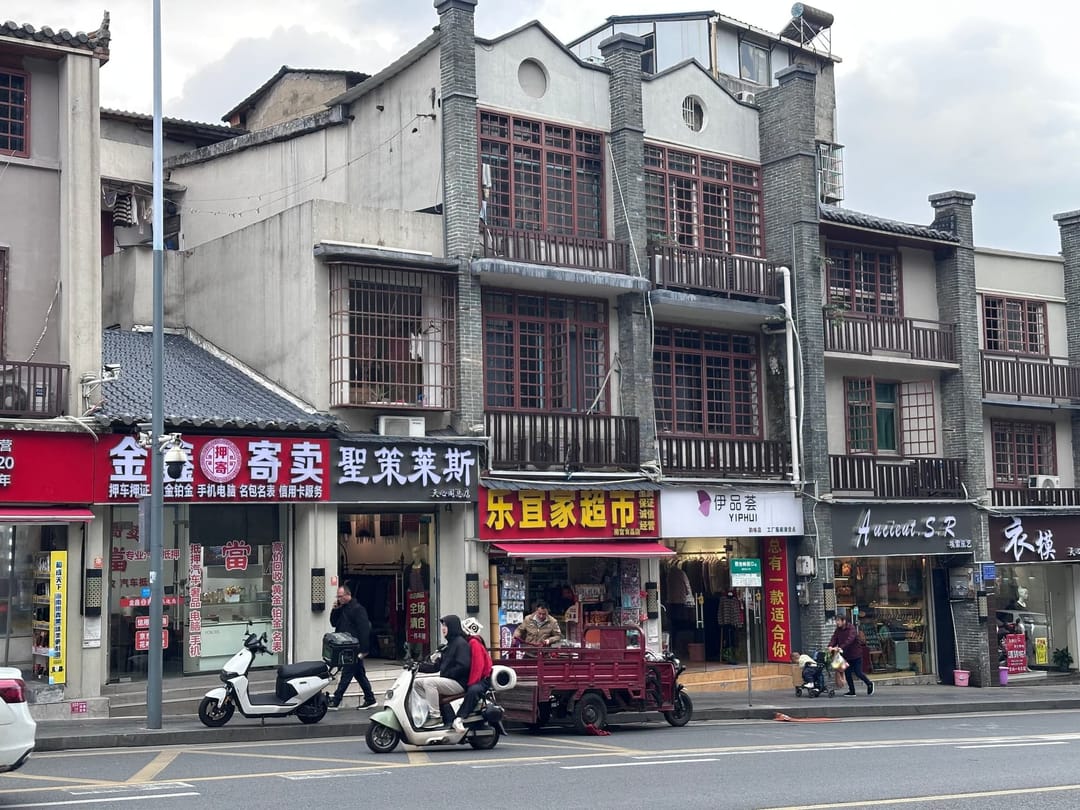 Rue commerçante de Changsha en journée : façades de brique grise à balcons de bois, enseignes rouges et jaunes en caractères, scooters et triporteur électrique au premier plan