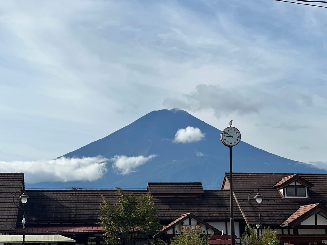 Le mont Fuji dégagé au-dessus d'une ceinture de nuages, vu depuis Kawaguchiko, toits d'une gare au premier plan avec une horloge