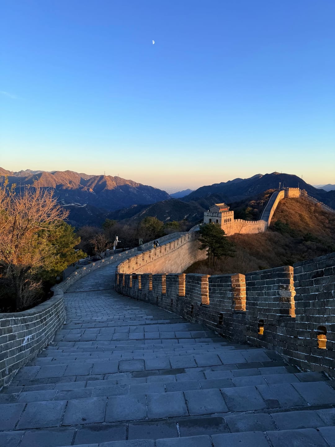 La Grande Muraille déserte à Badaling au coucher du soleil, parapets crénelés en ombre et muraille dorée serpentant vers les montagnes, lune visible
