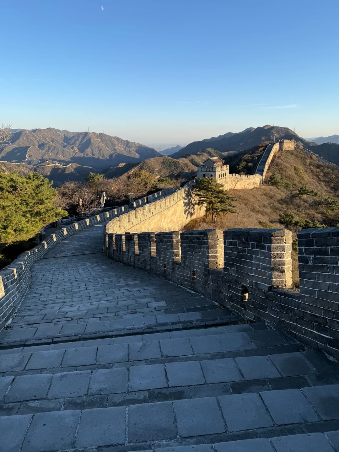 Chemin de ronde de la Grande Muraille à Badaling entièrement vide dans la lumière dorée du soir, tour de guet et crêtes au fond, lune pâle dans le ciel