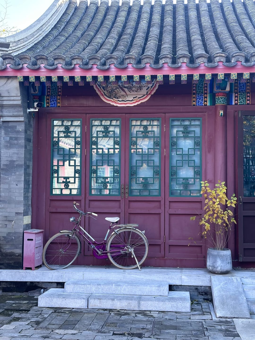 Façade repeinte d'une bâtisse de hutong : boiseries laquées lie-de-vin, fenêtres à claire-voie, toiture de tuiles grises, pavage neuf et un vélo appuyé devant les marches