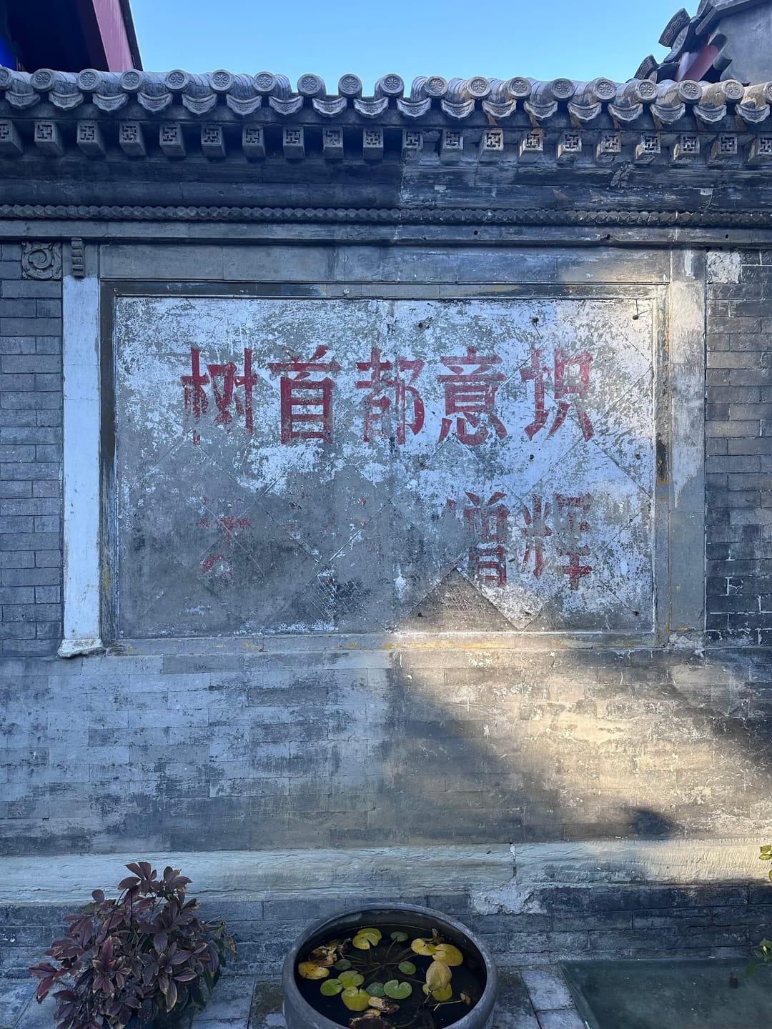 Mur-écran de hutong en brique grise coiffé de tuiles, portant de grands caractères rouges repeints, une jarre plantée à son pied