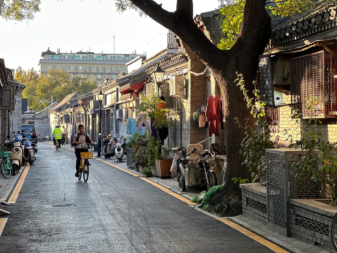 Ruelle résidentielle de hutong à Pékin bordée de grands arbres, vélos et scooters garés le long du trottoir, linge suspendu aux façades de brique, cycliste au loin