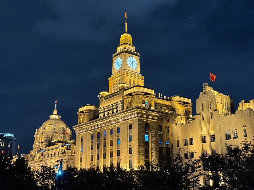 Deux bâtiments du Bund illuminés la nuit : à gauche la coupole de l'ancienne banque HSBC, à droite la tour-horloge de la Customs House, façades de pierre en lumière dorée sous un ciel bleu nuit
