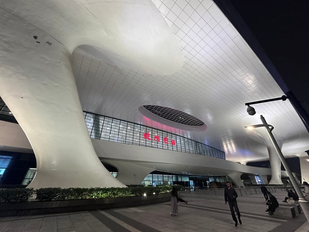 Façade et auvent incurvé blanc d'une gare à grande vitesse chinoise à la tombée du jour, panneaux d'information lumineux et voyageurs marchant vers l'entrée