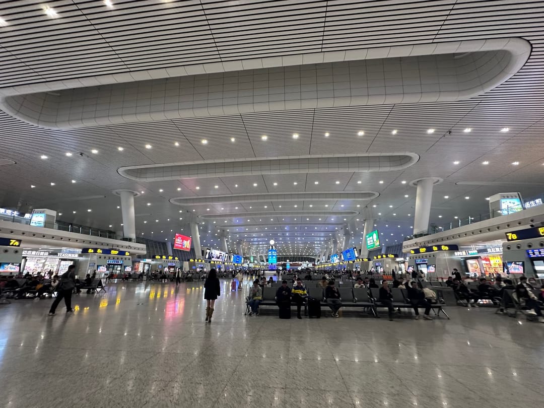 Hall d'attente d'une gare à grande vitesse chinoise, plafond nervuré, rangées de sièges occupés par des voyageurs avec leurs valises et commerces le long des murs