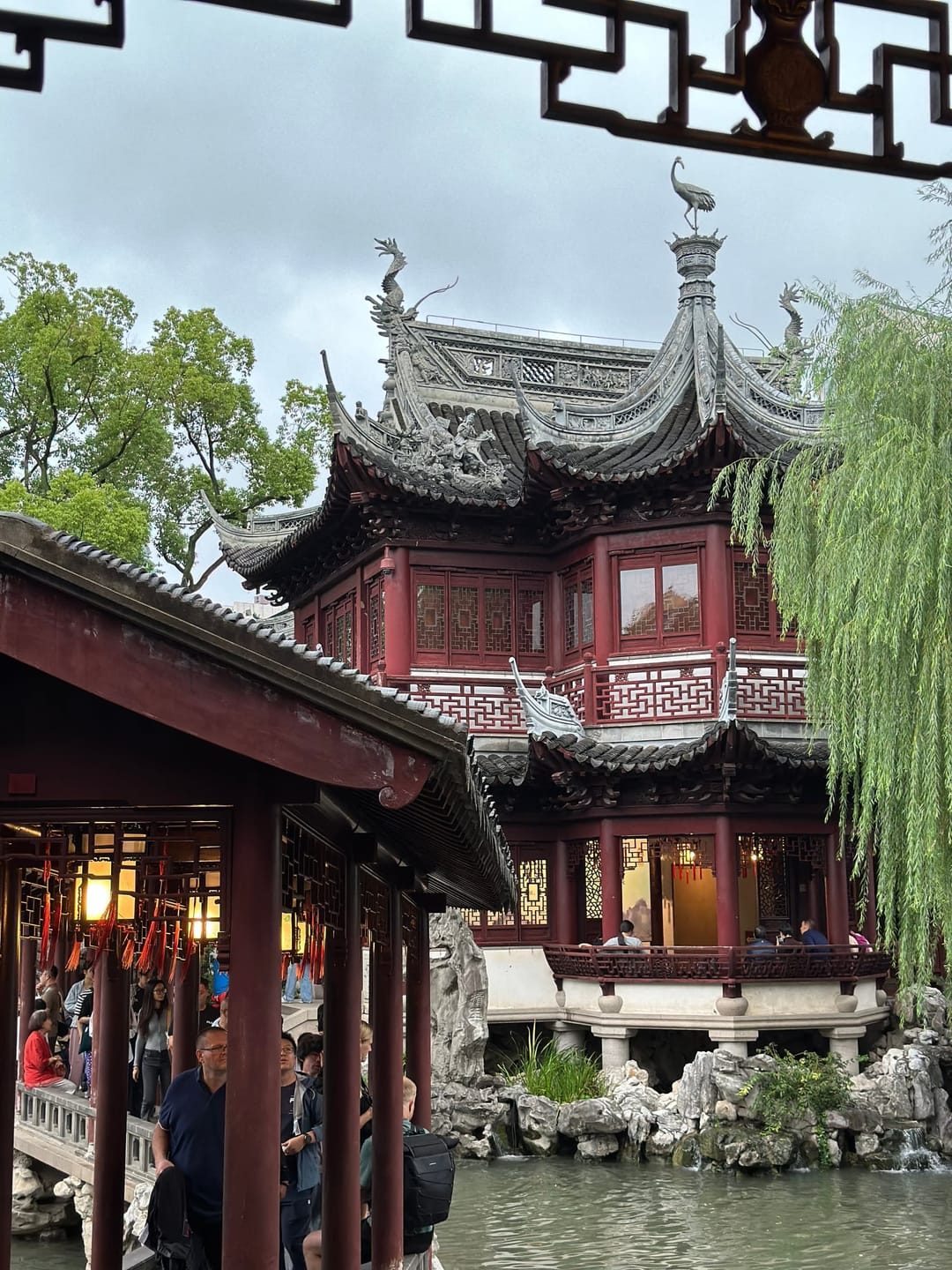 Pavillon à deux niveaux du jardin Yu surplombant un étang, boiseries sombres, saule pleureur et rocaille, visiteurs sous la galerie