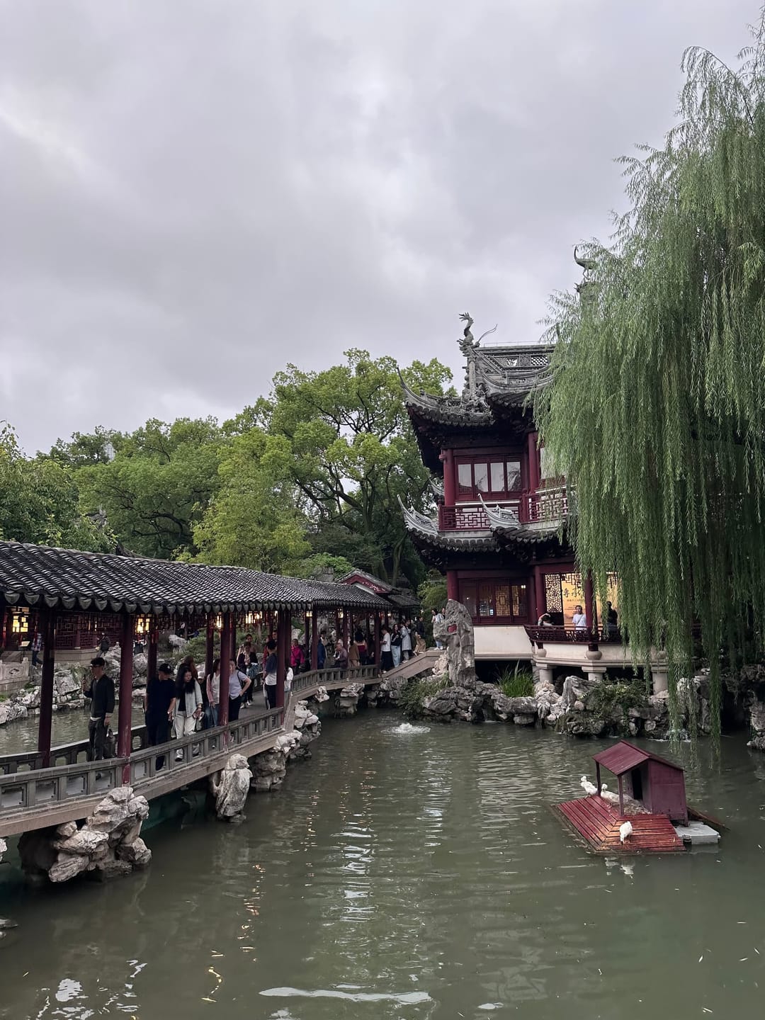 Galerie couverte longeant l'étang du jardin Yu, visiteurs alignés sous le toit, saules et barque rouge sur l'eau, ciel gris