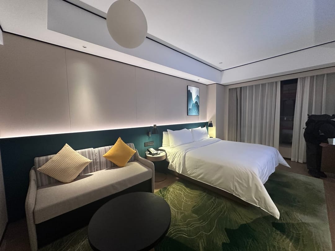 Chambre d'hôtel spacieuse en Chine : grand lit blanc, canapé et coussins jaunes, tapis vert, éclairage tamisé et estampe de montagne au mur