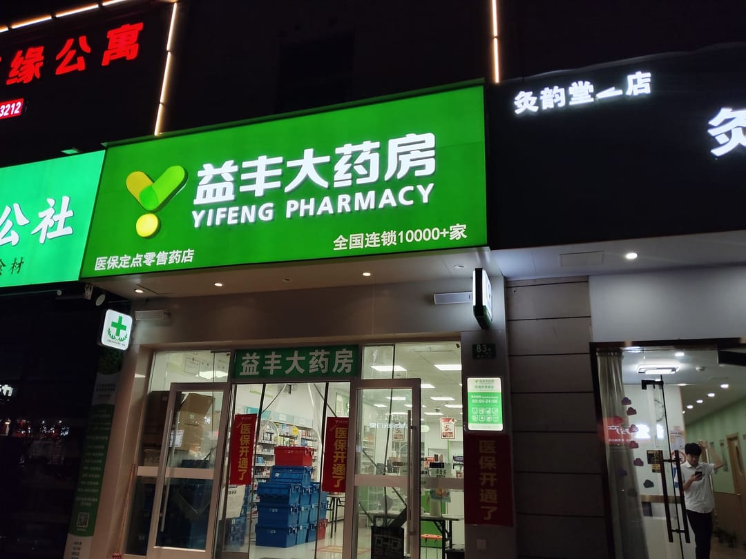 Devanture éclairée d'une pharmacie Yifeng à Shanghai, la nuit : enseigne verte bilingue avec croix de pharmacie, portes vitrées ouvertes sur des rayonnages éclairés