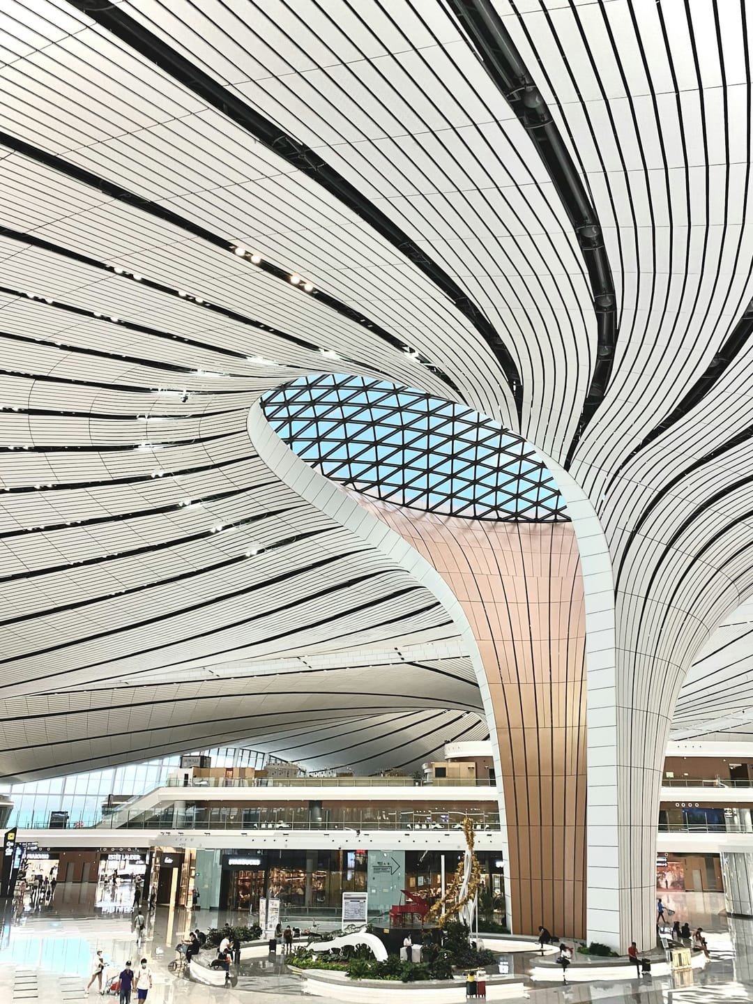 Hall de l'aéroport de Pékin Daxing, immense toiture blanche en éventail soutenue par des piliers évasés et percée d'un puits de lumière, voyageurs minuscules au sol