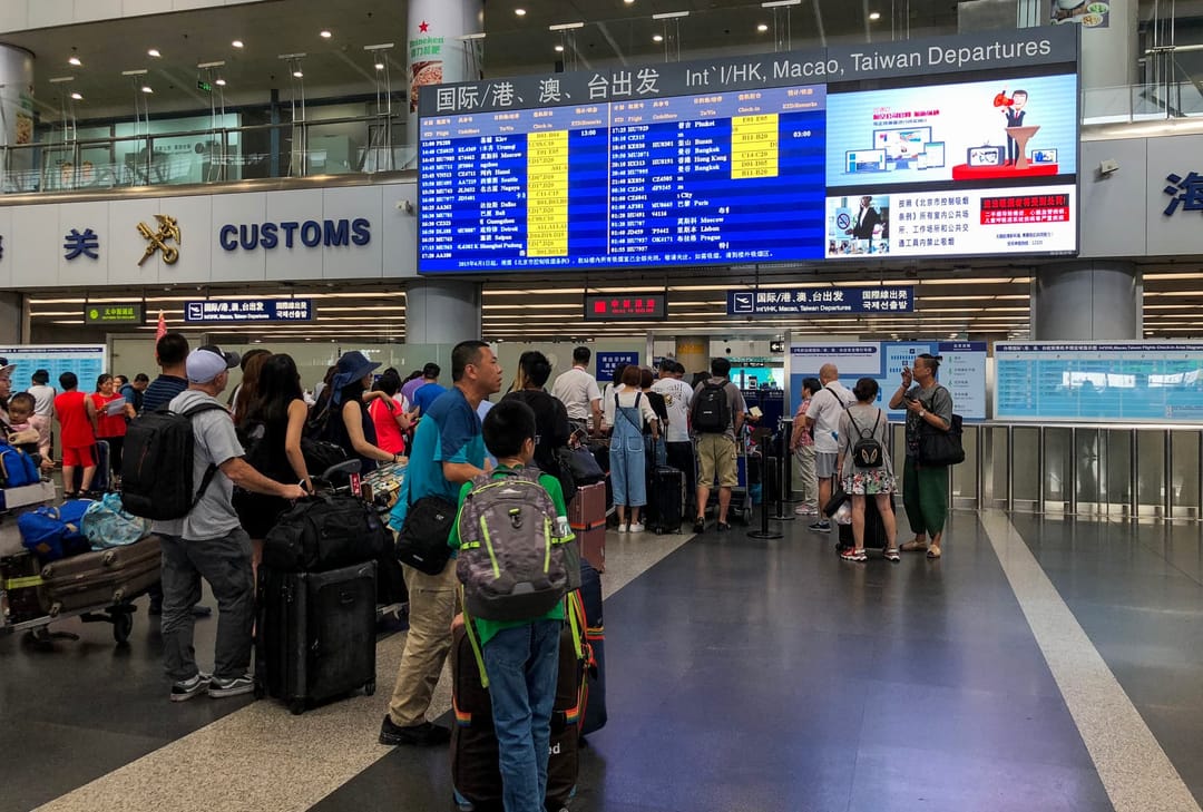 Hall des départs internationaux du terminal 2 de l'aéroport de Pékin : une file de voyageurs devant les comptoirs d'enregistrement, valises à roulettes et sacs à dos au sol, écran d'affichage des vols bilingue au-dessus et panneau Customs à gauche
