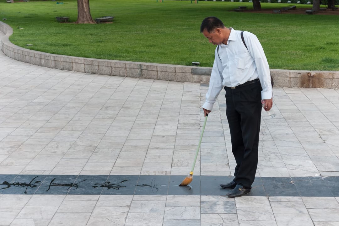 Un homme trace des caractères à l'eau sur le dallage d'un parc de Dalian, penché sur un long pinceau-éponge, une bouteille d'eau dans l'autre main, les caractères déjà tracés s'effaçant derrière lui