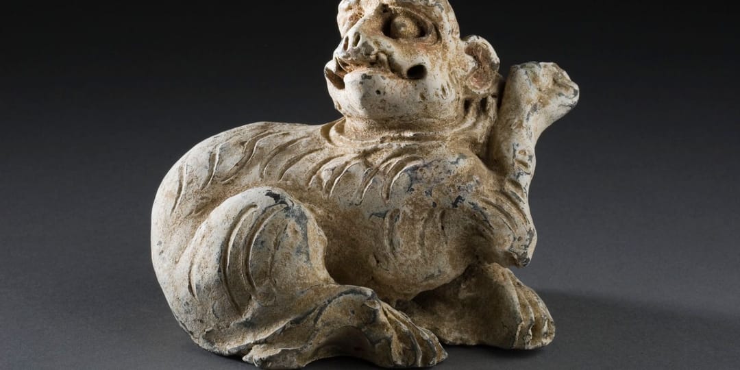Figurine funéraire chinoise en terre cuite beige représentant un animal couché aux traits de félin, la tête relevée et la patte avant dressée, photographiée sur un fond gris neutre de musée