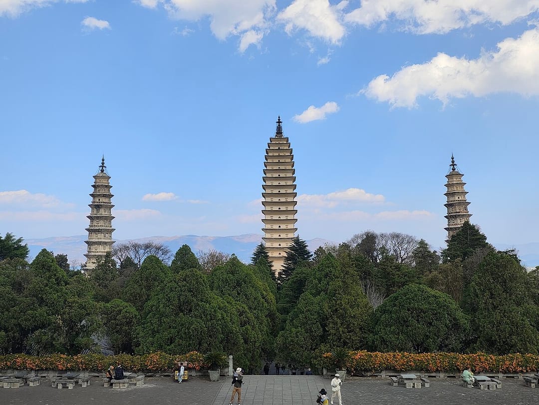 Les trois pagodes de Chongsheng sous un ciel bleu : au centre la haute pagode Qianxun à seize étages de briques claires, encadrée par deux pagodes plus petites et plus trapues, au-dessus d'une masse de conifères sombres et d'une esplanade dallée