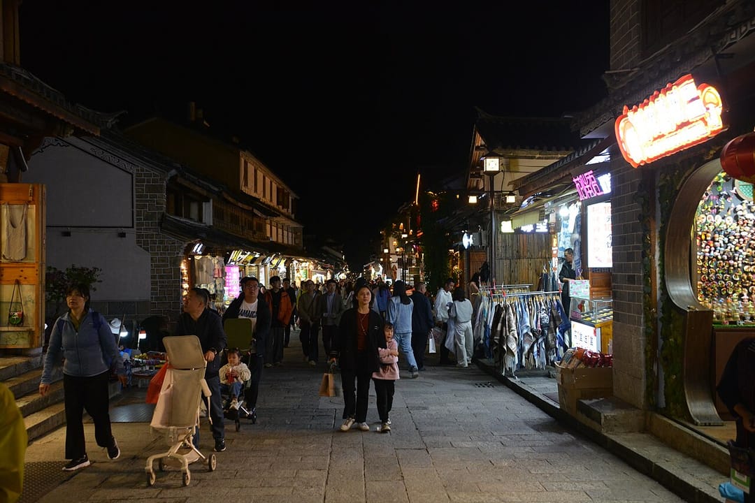 Rue pavée de la vieille ville de Dali le soir, bordée de façades en briques grises et bois sombre, avec des enseignes lumineuses, des étals de vêtements et des passants dont une famille avec une poussette