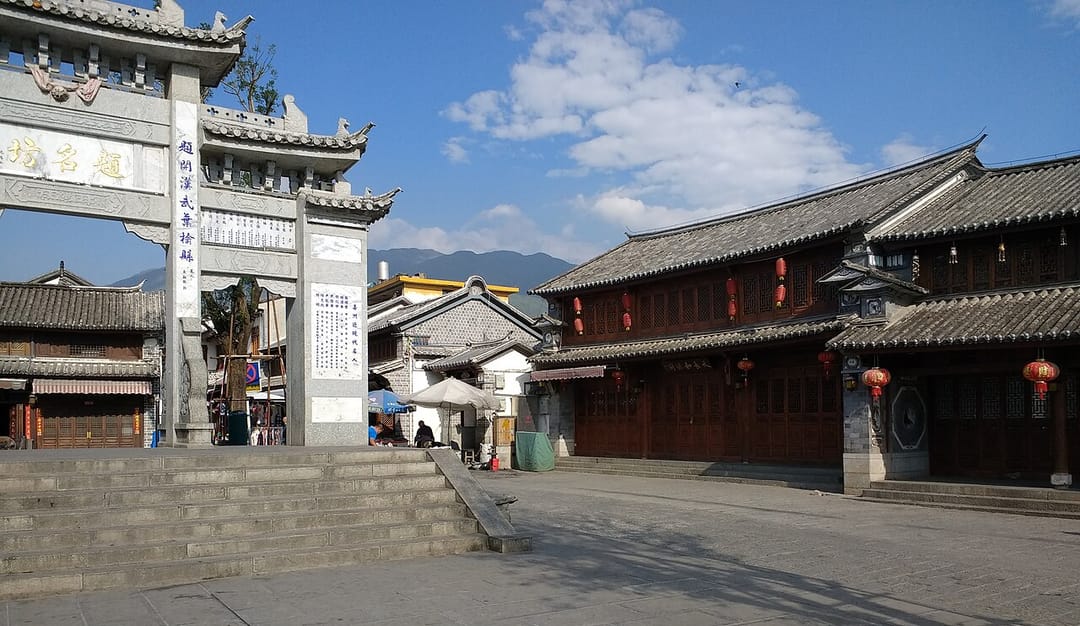 Place de Xizhou : à gauche une haute porte monumentale en pierre grise couverte d'inscriptions chinoises, à droite un bâtiment bai à façade de bois sombre ajouré et lanternes rouges sous un toit de tuiles grises, avec la montagne en arrière-plan