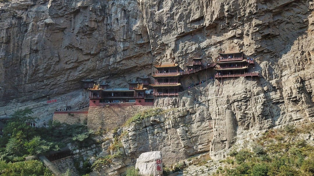 Le monastère suspendu de Xuankong accroché à une falaise du mont Heng : pavillons de bois rouge sur pilotis étagés le long de la paroi, passerelles reliant les bâtiments, visiteurs minuscules sur les coursives